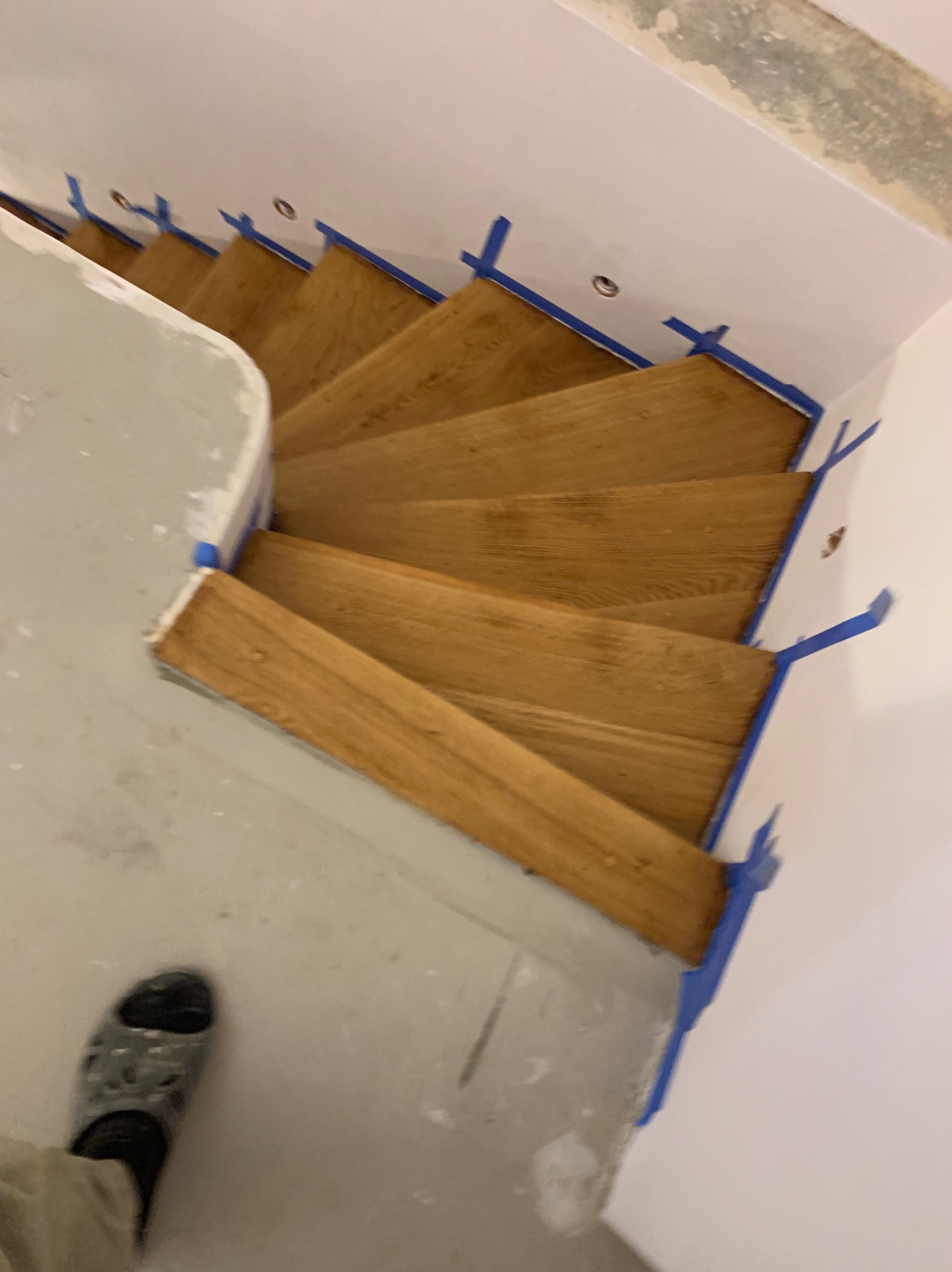 Drewniane schody po cyklinowaniu, zabezpieczone taśmą malarską, widok z góry. Stopa w klapku w lewym dolnym rogu kadru. Jasne wnętrze w trakcie prac wykończeniowych.