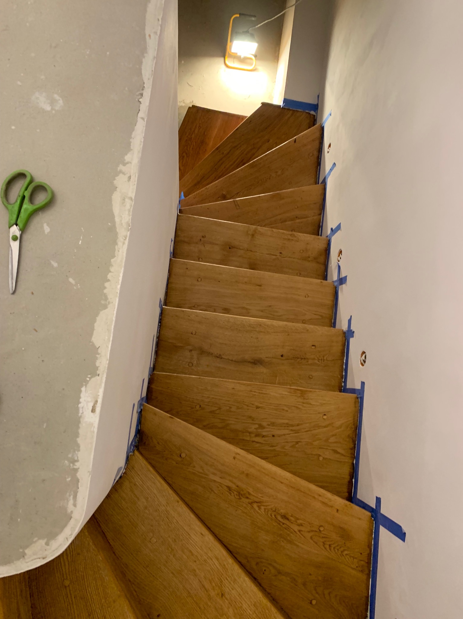 Drewniane schody spiralne w trakcie montażu, zabezpieczone niebieską taśmą malarską, widoczne nożyczki na szarym podłożu. Górne oświetlenie.