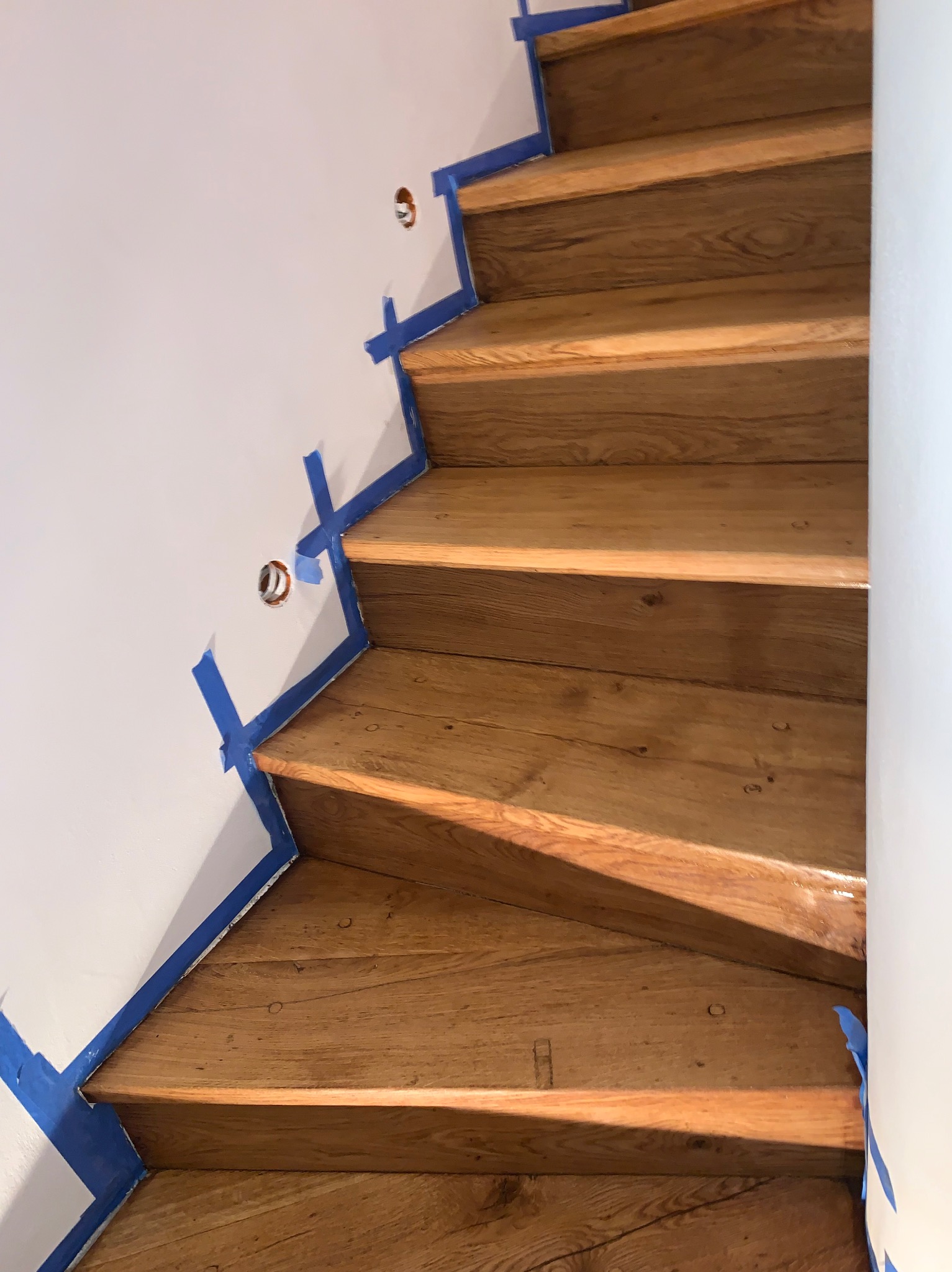 Odnowione drewniane schody z zabezpieczającą niebieską taśmą malarską przy białej ścianie. Widok z góry na błyszczące, polakierowane stopnie.