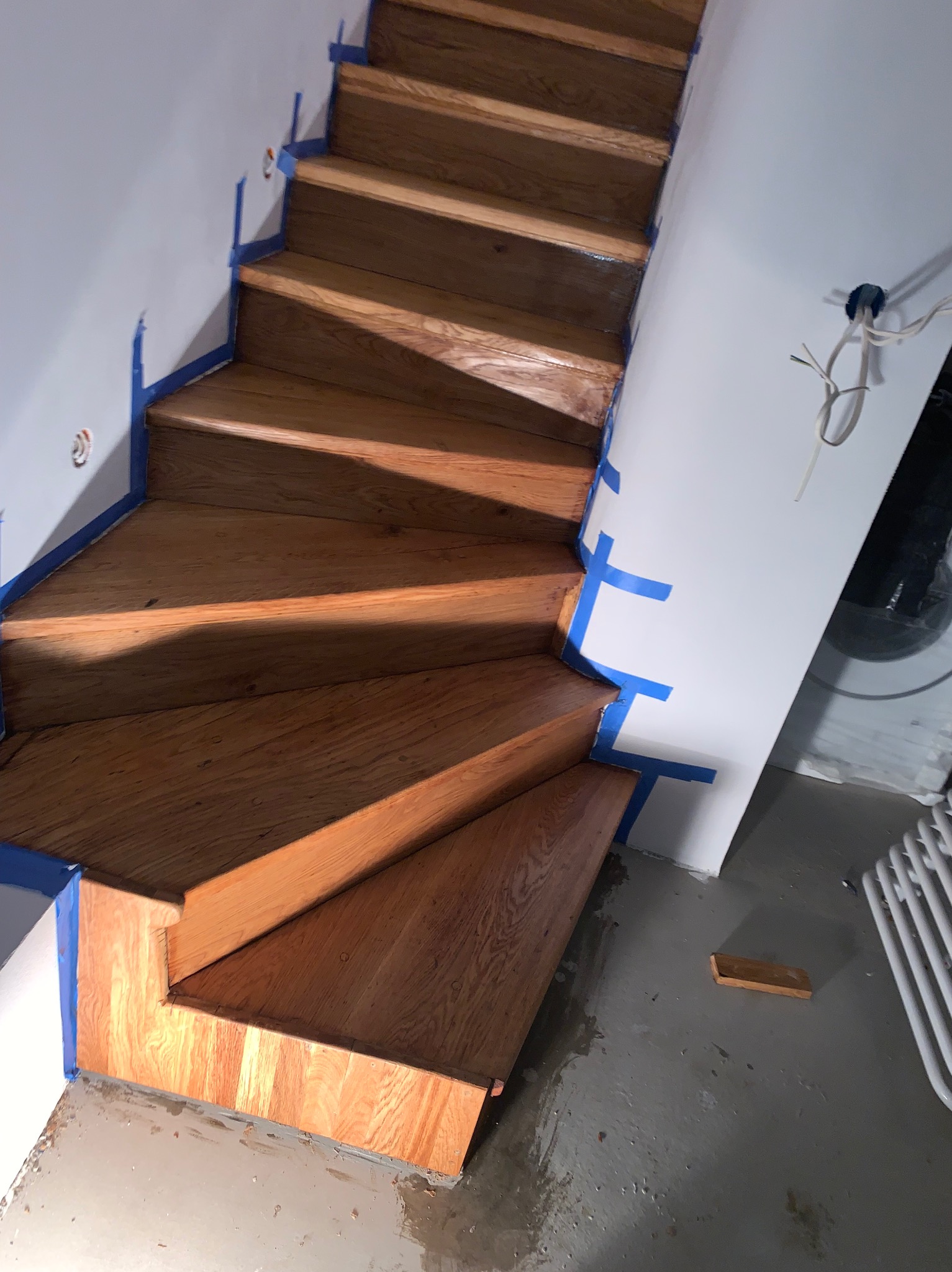 Drewniane schody po renowacji, zabezpieczone niebieską taśmą malarską. Widoczne świeżo polakierowane stopnie i fragment ściany z puszką elektryczną.