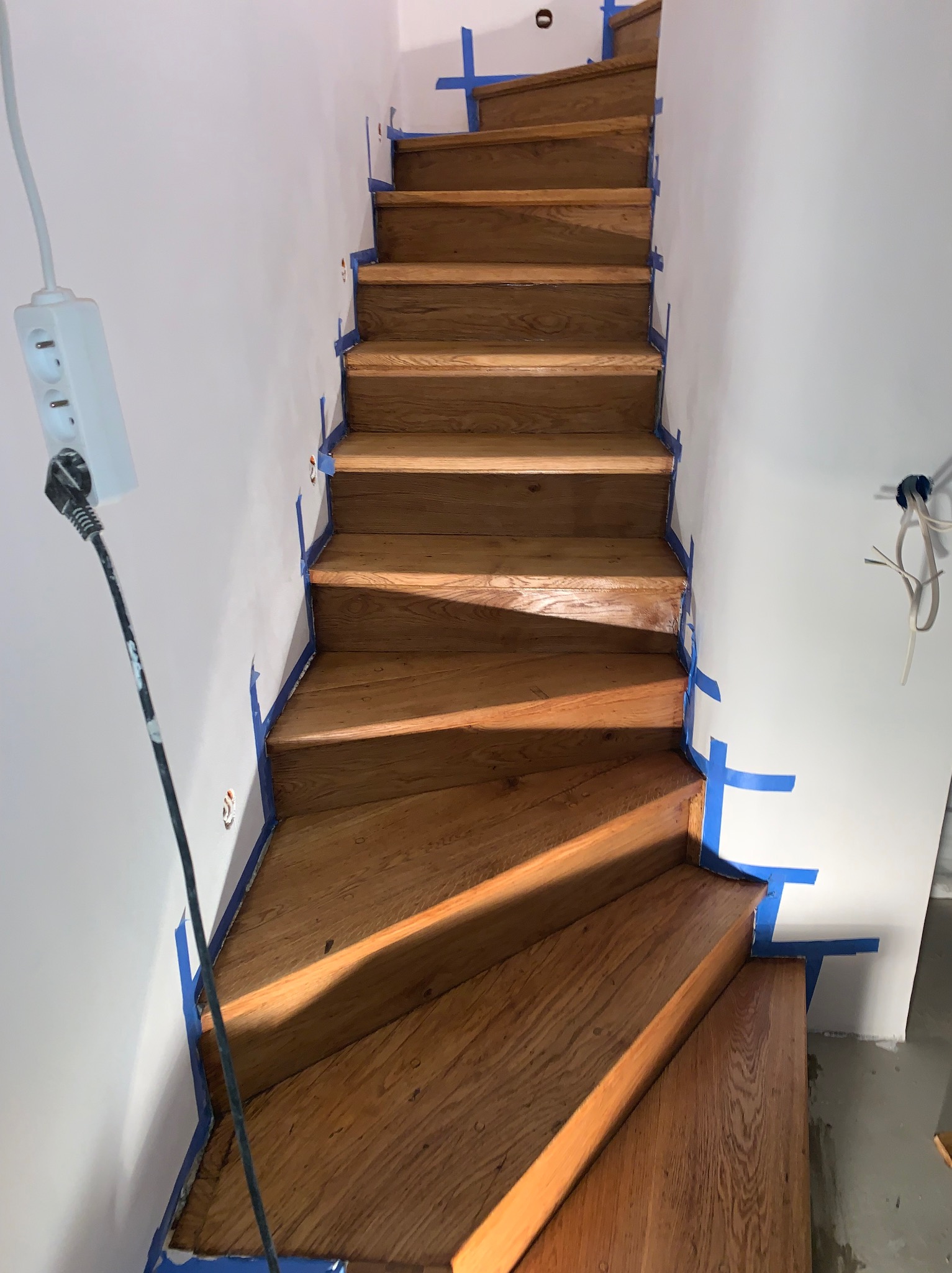 Drewniane schody po renowacji, zabezpieczone niebieską taśmą malarską przy ścianach. Widoczne gniazdko elektryczne i przewody. Jasne, naturalne oświetlenie.