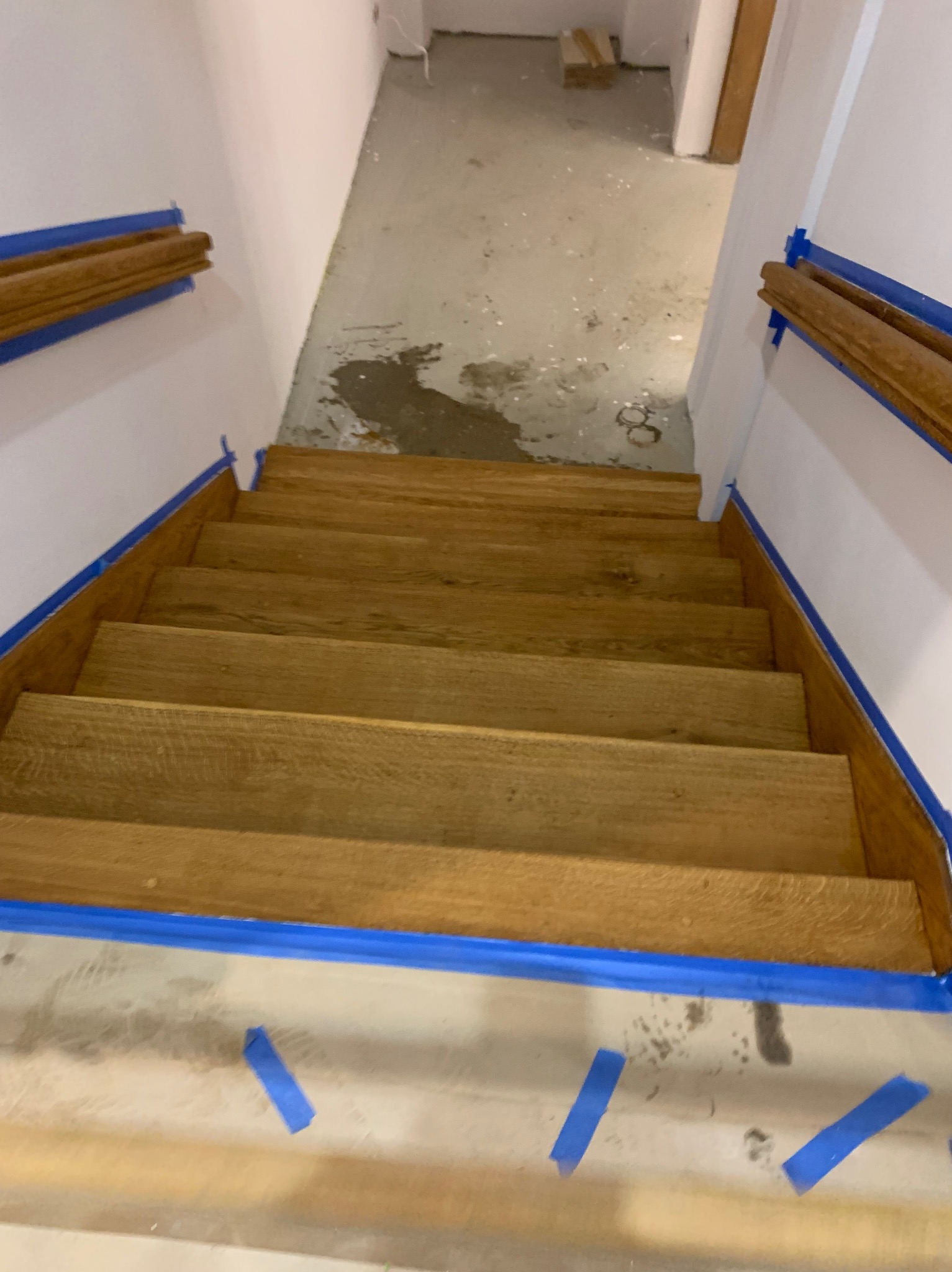 Drewniane schody po renowacji, zabezpieczone niebieską taśmą malarską. Widok z góry, jasne oświetlenie, fragment betonowej podłogi w tle.