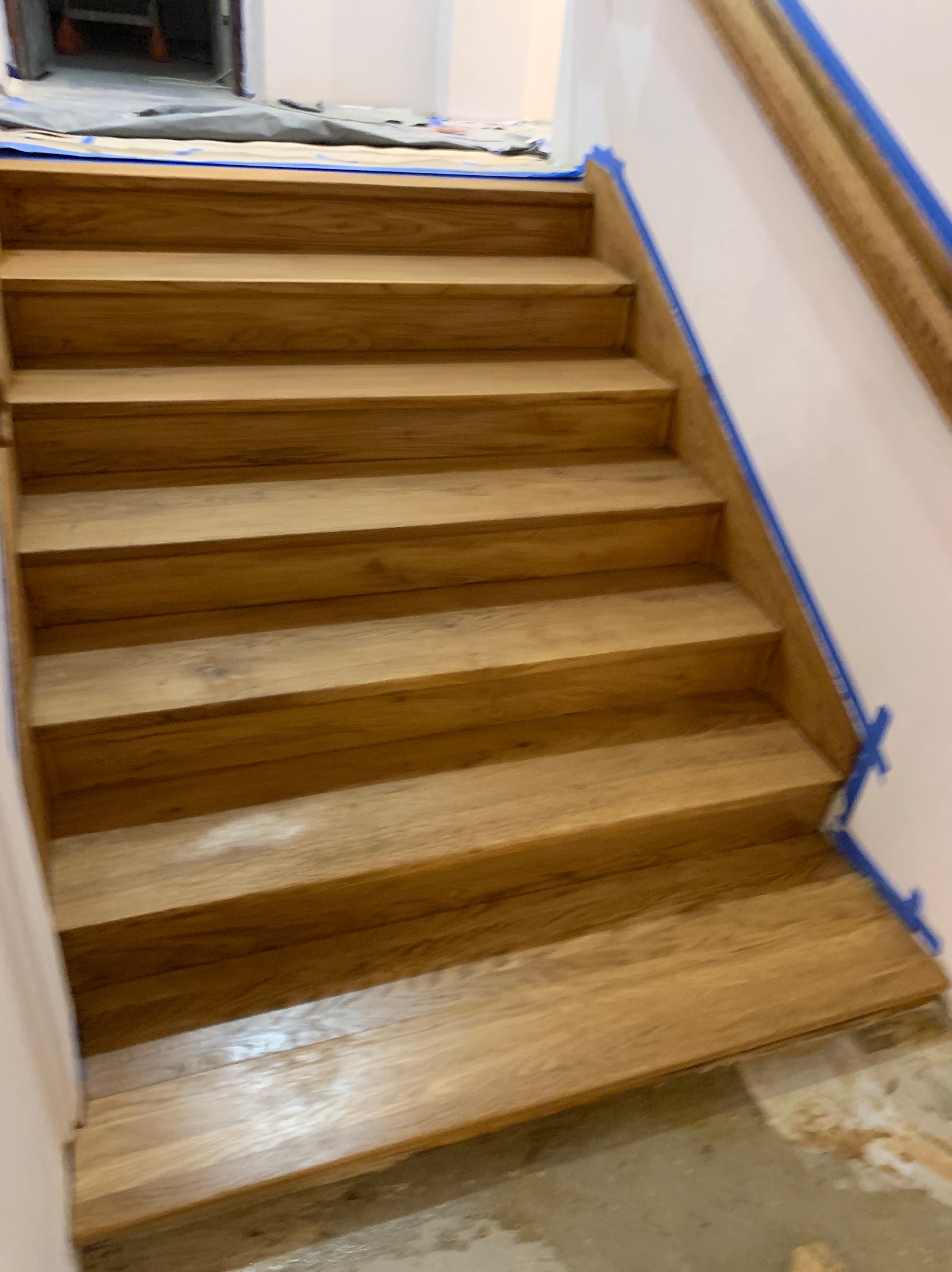 Odnowione drewniane schody z zabezpieczonymi taśmą malarską ścianami. Widoczne detale słojów drewna i świeżo położony lakier. Ujęcie z dołu schodów.
