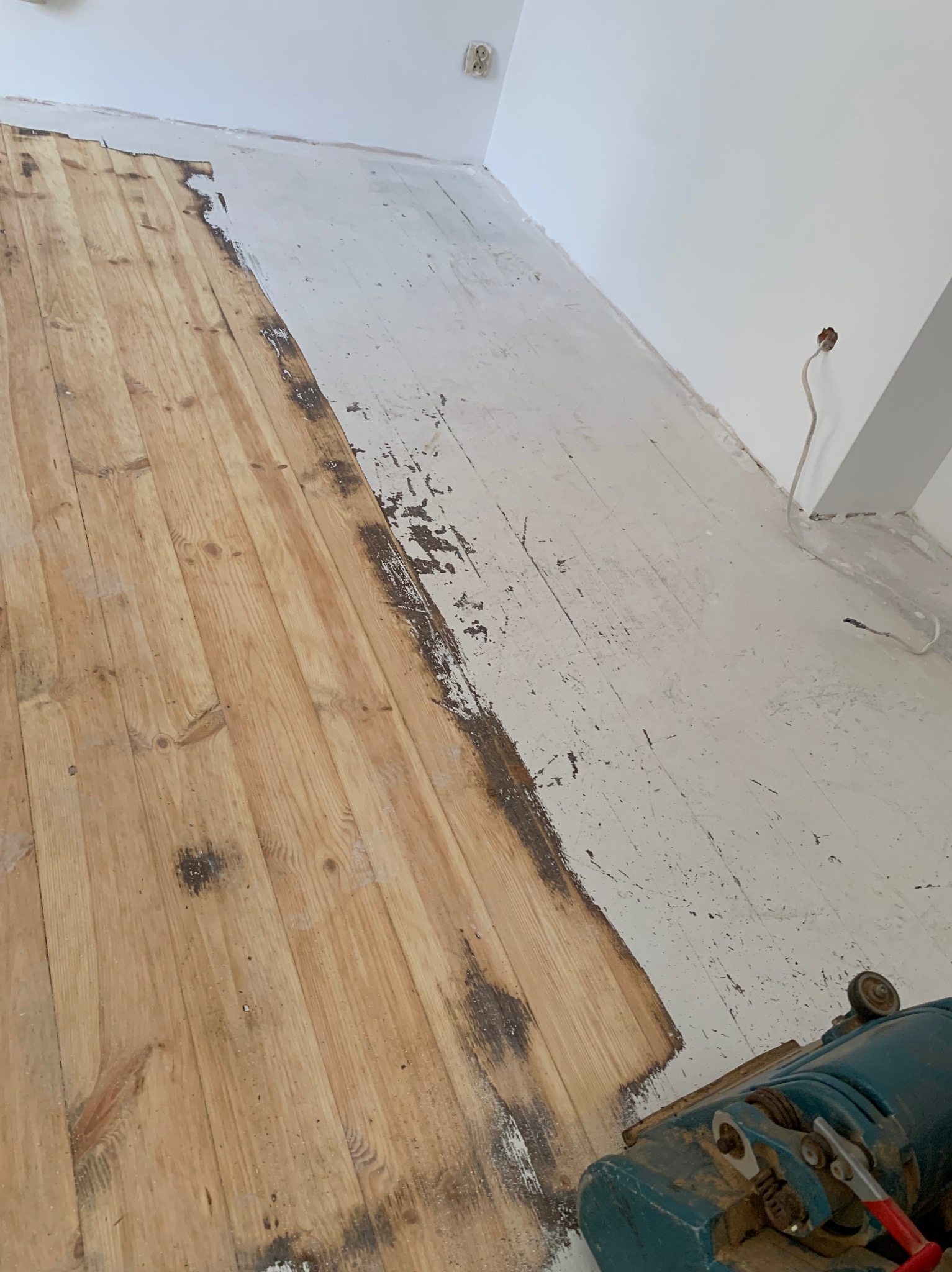 Częściowo odnowiona podłoga drewniana z widoczną maszyną do cyklinowania. Połowa podłogi w naturalnym kolorze drewna, druga połowa z resztkami starej farby.