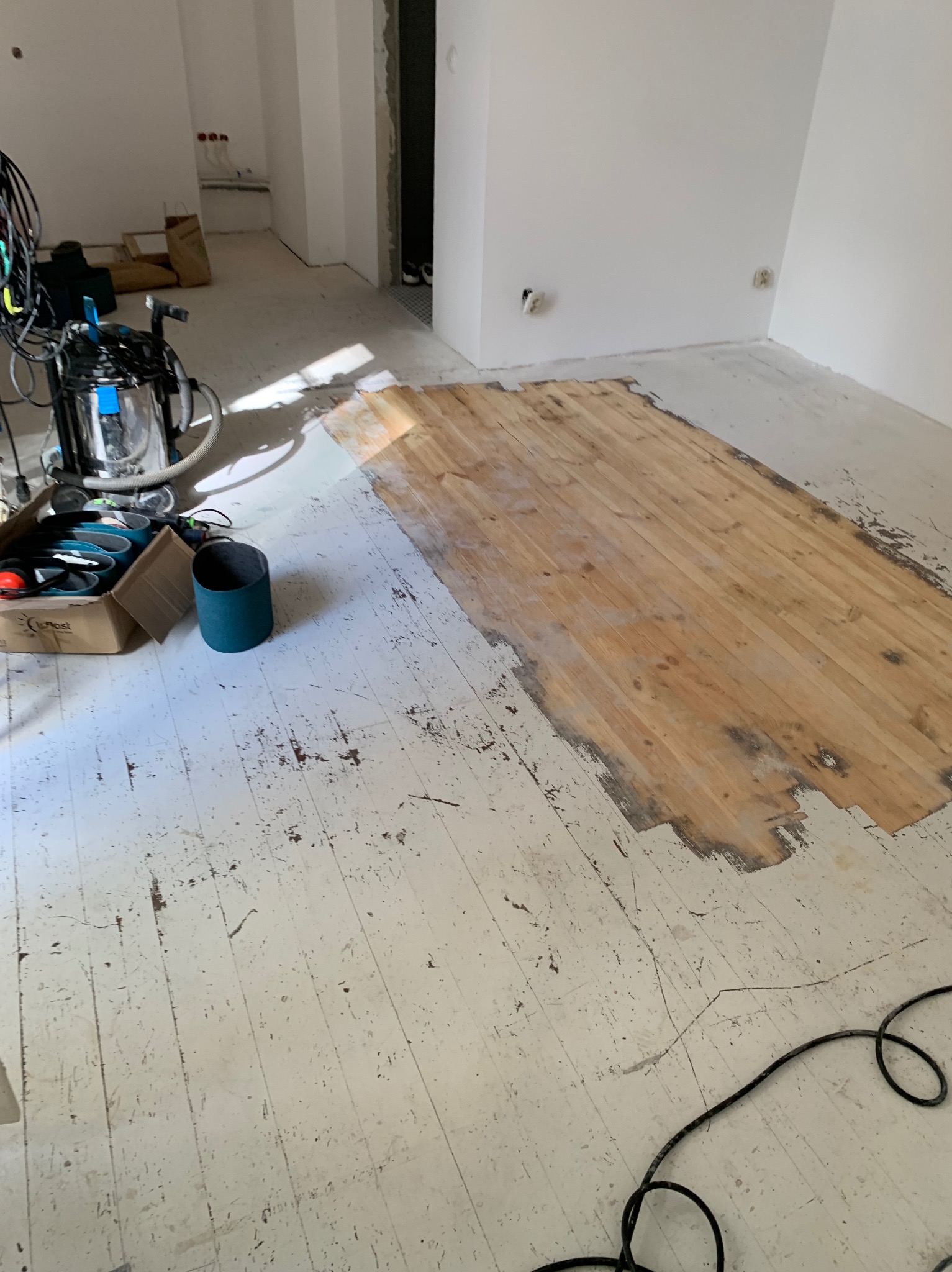 Częściowo wycyklinowana podłoga drewniana w jasnym pomieszczeniu. Widoczna maszyna do cyklinowania, akcesoria i fragmenty starej farby na podłodze.
