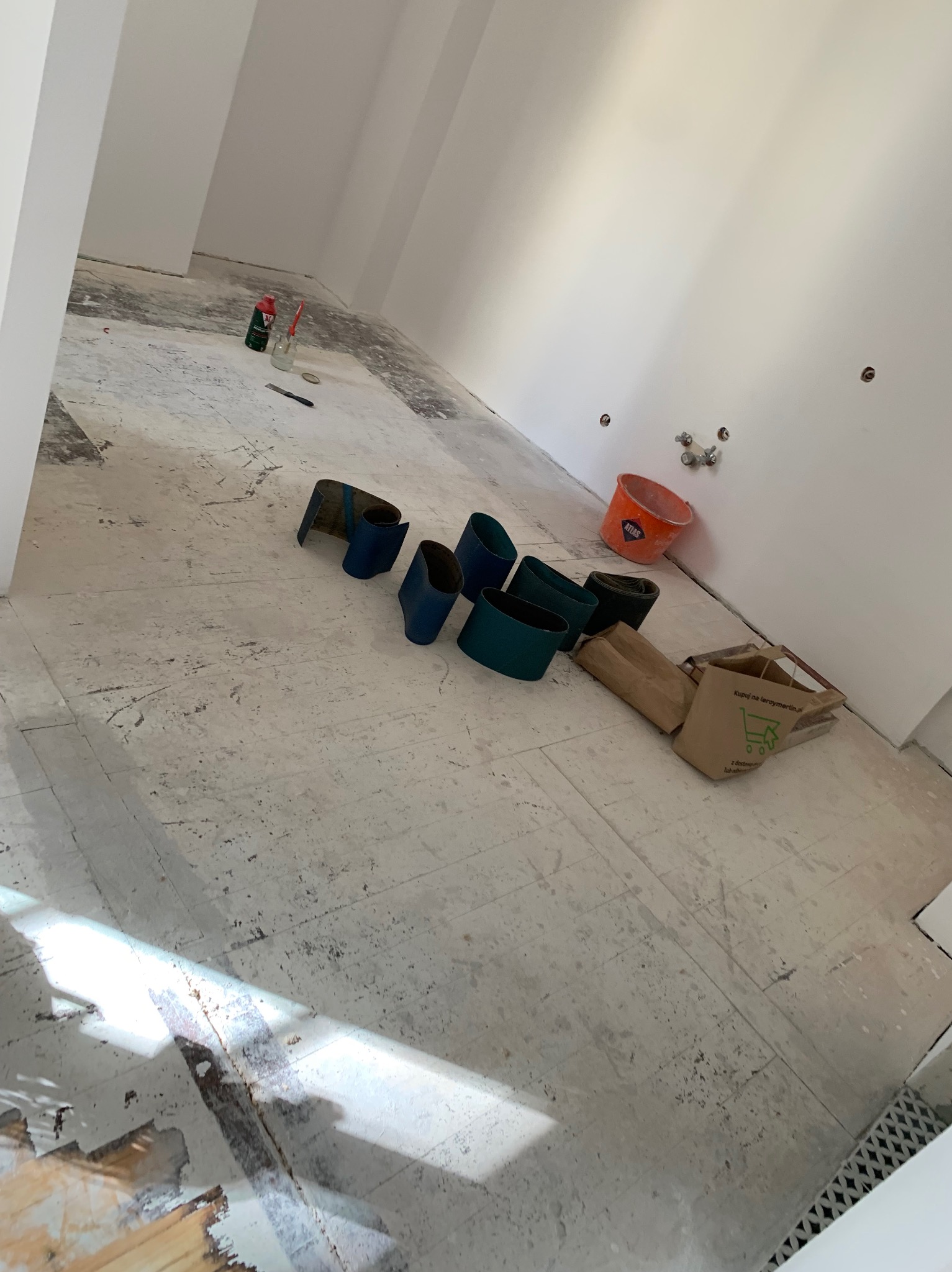 Przygotowanie do cyklinowania: zmatowiona podłoga, wałki ścierne, wiadro i torba na zakupy w pustym, jasnym pomieszczeniu. Widoczne ślady po starej powłoce.