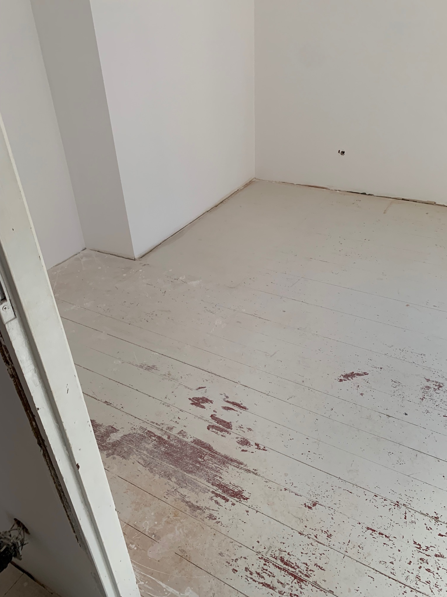 Biała podłoga z desek z widocznymi śladami starej farby i ubytkami, fragment ściany i futryny drzwi. Widok na odnawianą podłogę w pustym pomieszczeniu.