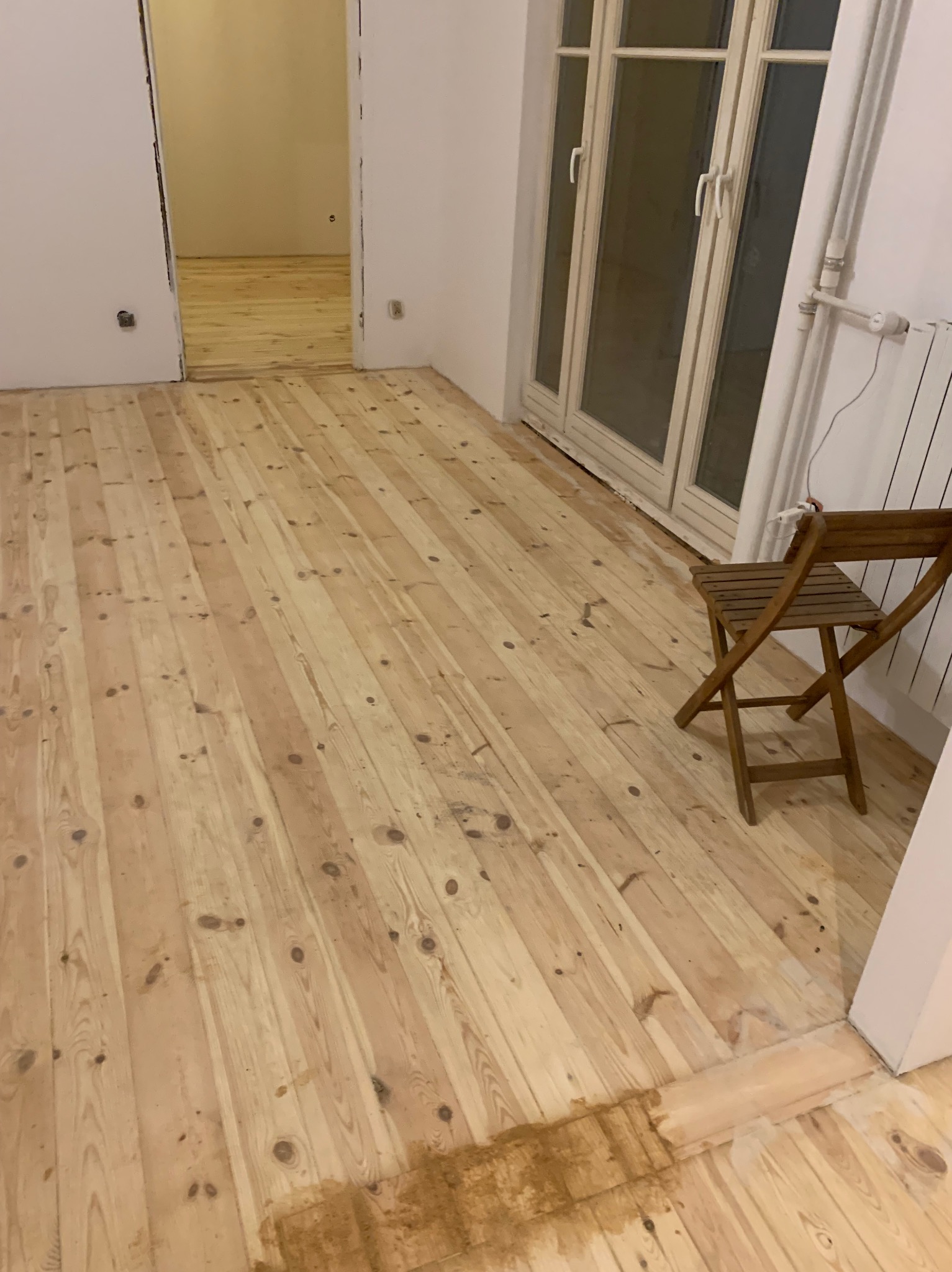 Jasna, odnowiona podłoga z desek w pokoju z białymi ścianami i oknem. Na podłodze stoi drewniane krzesło. Widoczny fragment podłogi w sąsiednim pomieszczeniu.