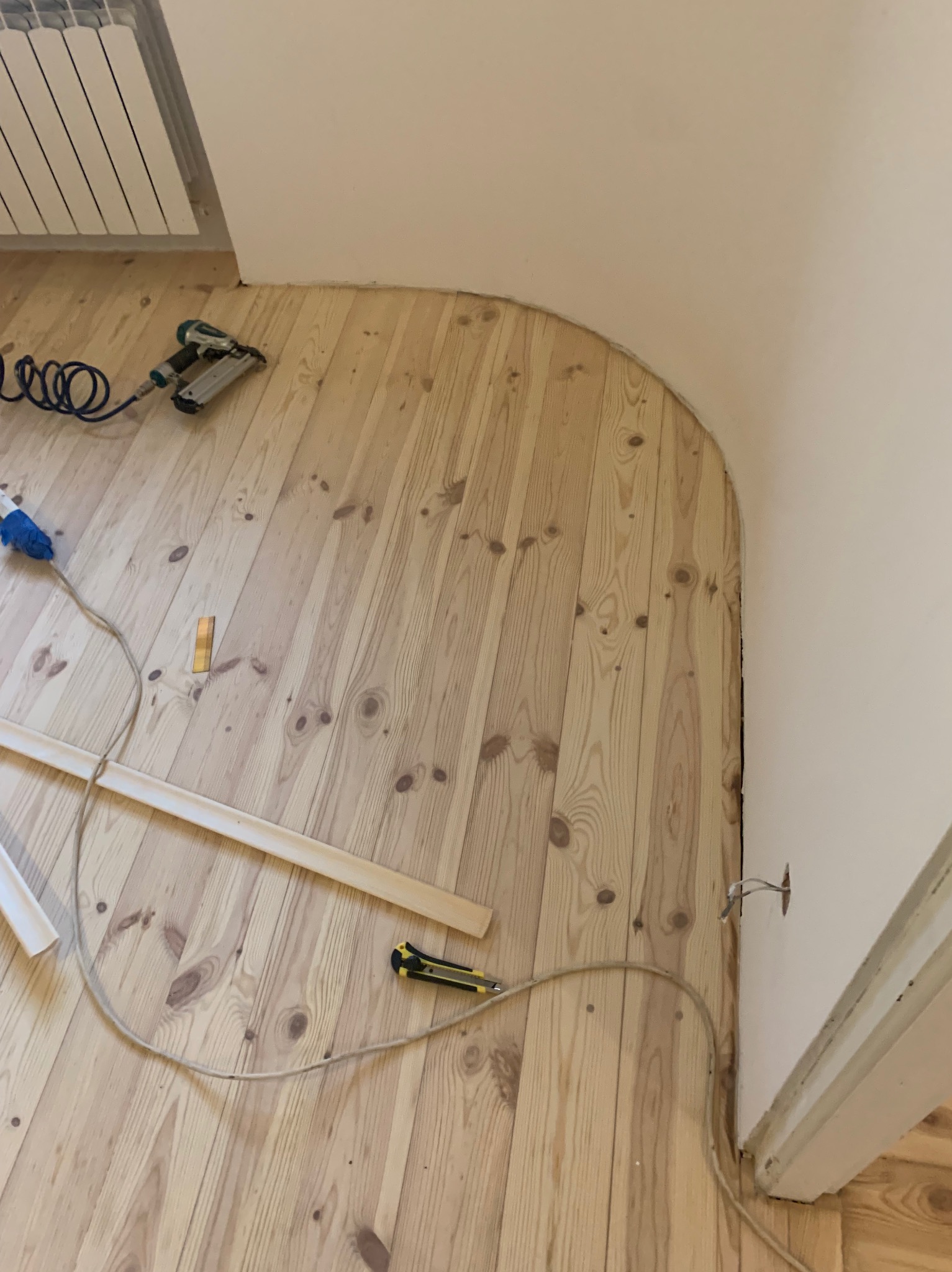 Jasna drewniana podłoga z narzędziami do montażu listew przypodłogowych przy zaokrąglonej ścianie. Widoczny zszywacz pneumatyczny i nóż do cięcia.