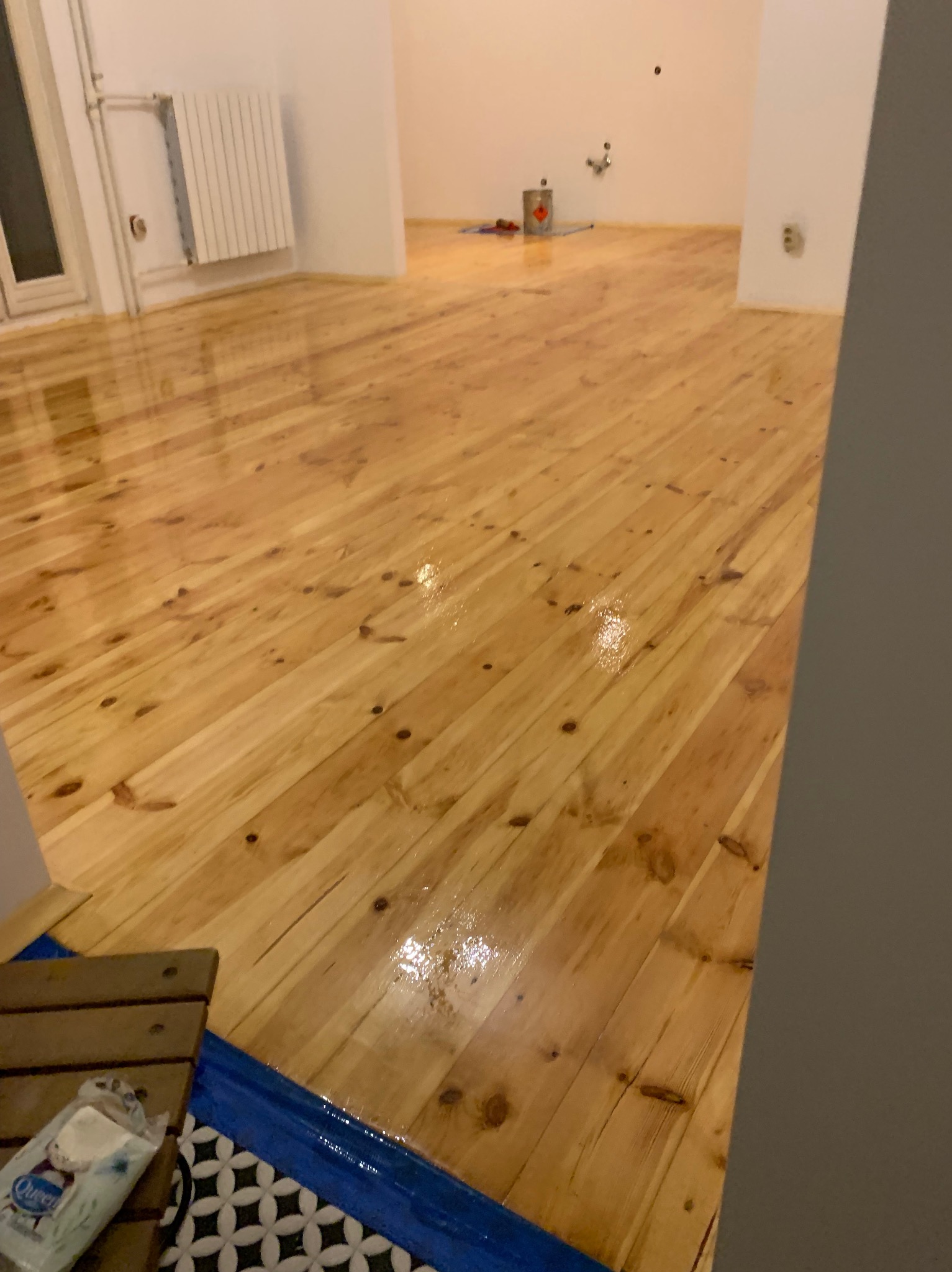 Błyszcząca, świeżo polakierowana podłoga z jasnego drewna w pustym pokoju. Widoczne ślady lakieru i odbicia światła. W tle grzejnik i puszka z lakierem.