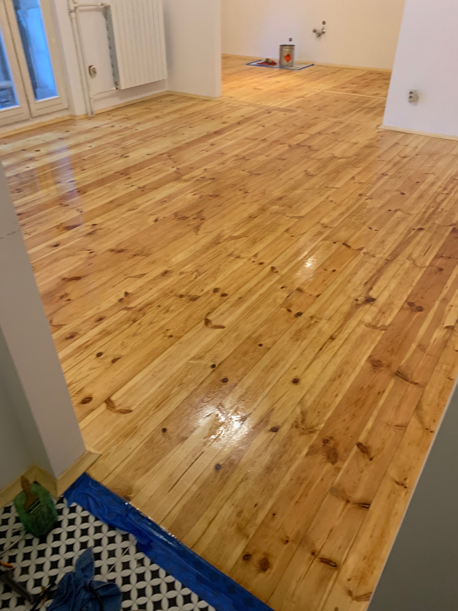 Błyszcząca, świeżo polakierowana podłoga z desek w odcieniach miodowego brązu. Widoczne zabezpieczenia folią malarską przy ścianie i puszka z lakierem w tle.