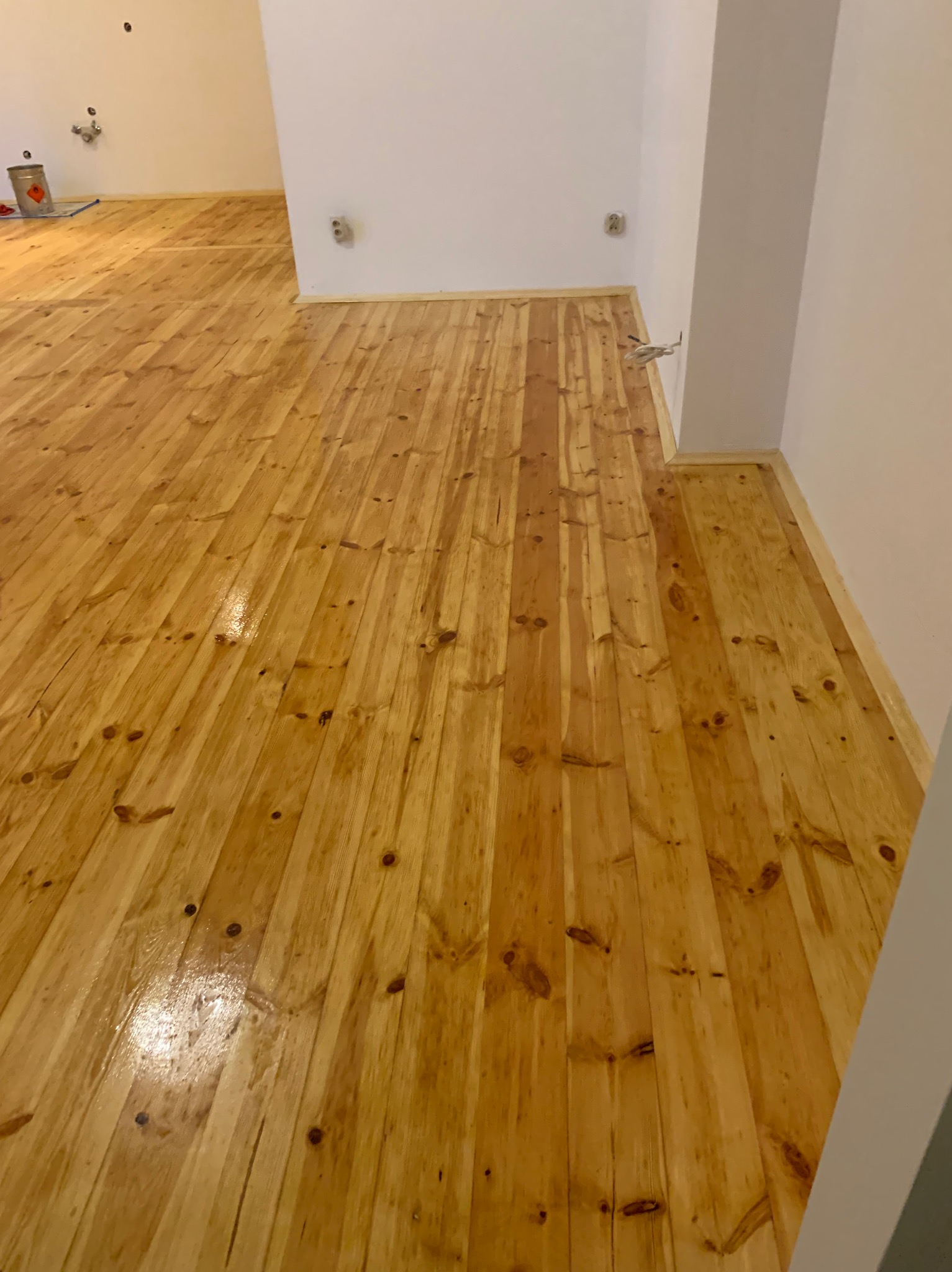 Błyszcząca, świeżo polakierowana podłoga z jasnego drewna w pustym pokoju. Widoczne ślady lakieru i naturalne usłojenie drewna. W tle ściana z wystającymi elementami instalacji.
