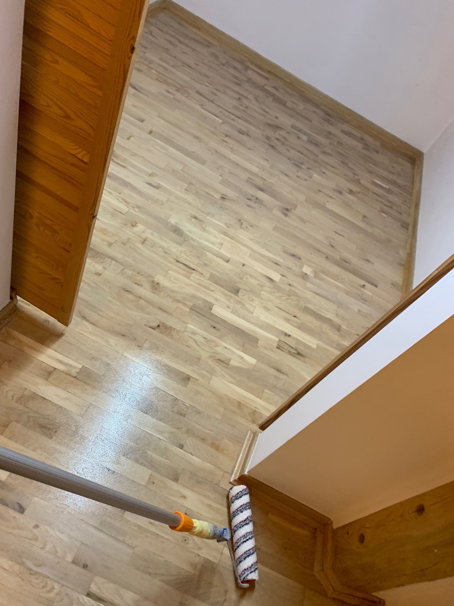 Lakierowanie parkietu wałkiem z długim uchwytem. Jasne drewno, widoczne słoje. Fragment ściany i drzwi w tle. Ujęcie z góry, ukośne.