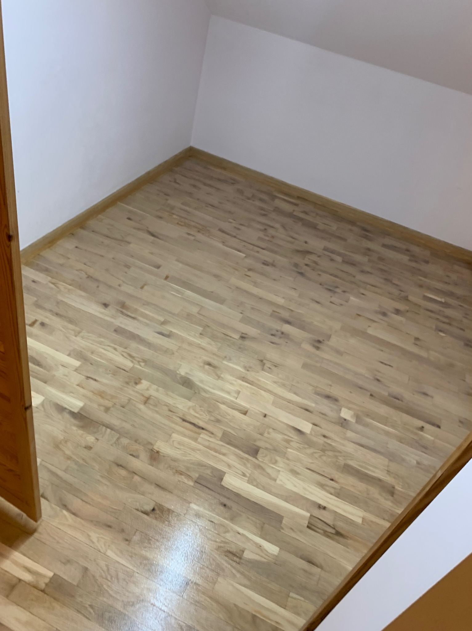 Jasna, drewniana podłoga parkietowa ułożona w nowym pomieszczeniu z białymi ścianami. Widoczny fragment ościeżnicy drzwi. Parkiet świeżo po cyklinowaniu, z połyskiem.