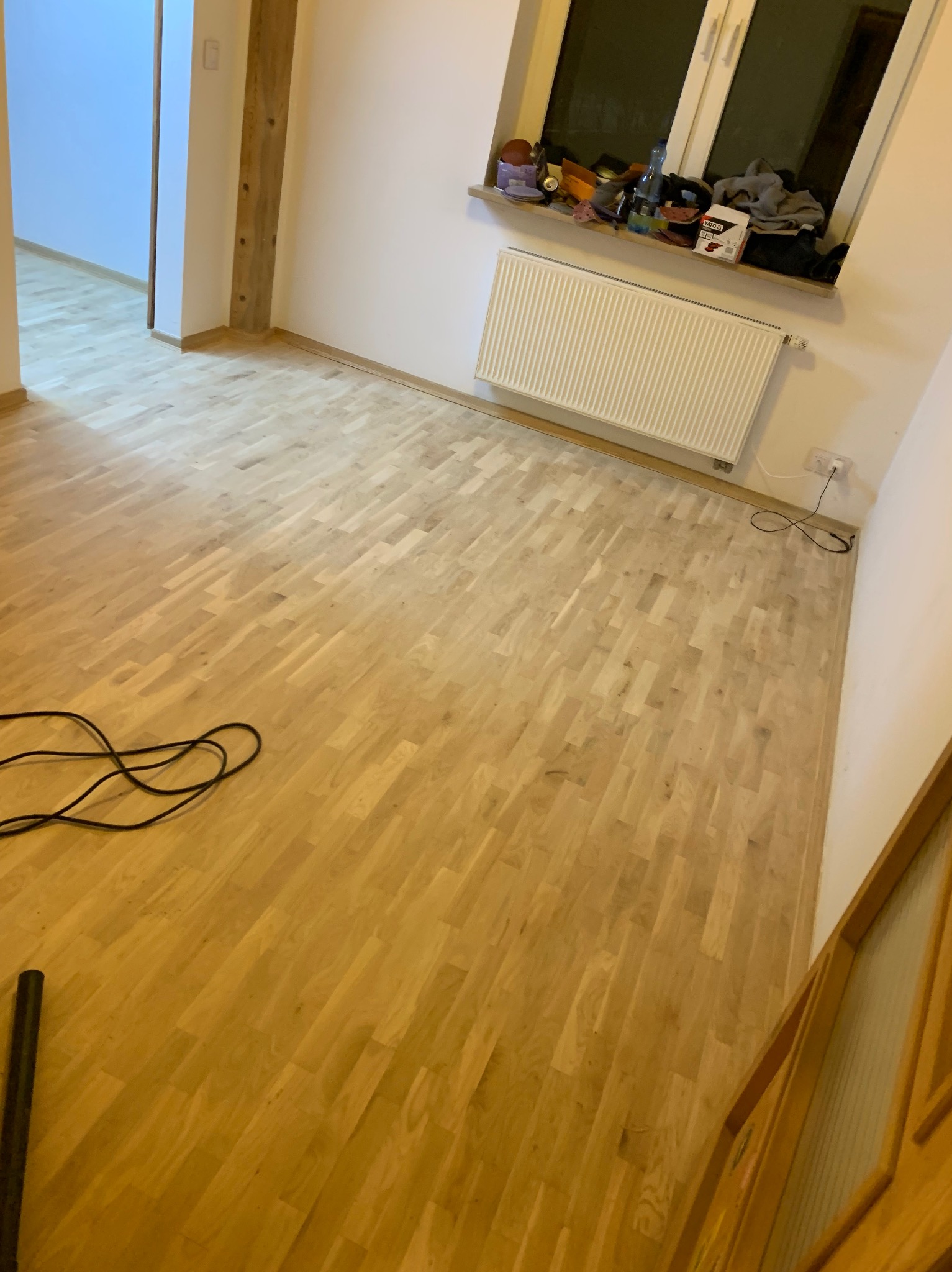 Jasna, drewniana podłoga parkietowa w pokoju z grzejnikiem pod oknem. Widoczny kabel i fragment rury. Świeżo położony parkiet w nowym mieszkaniu.