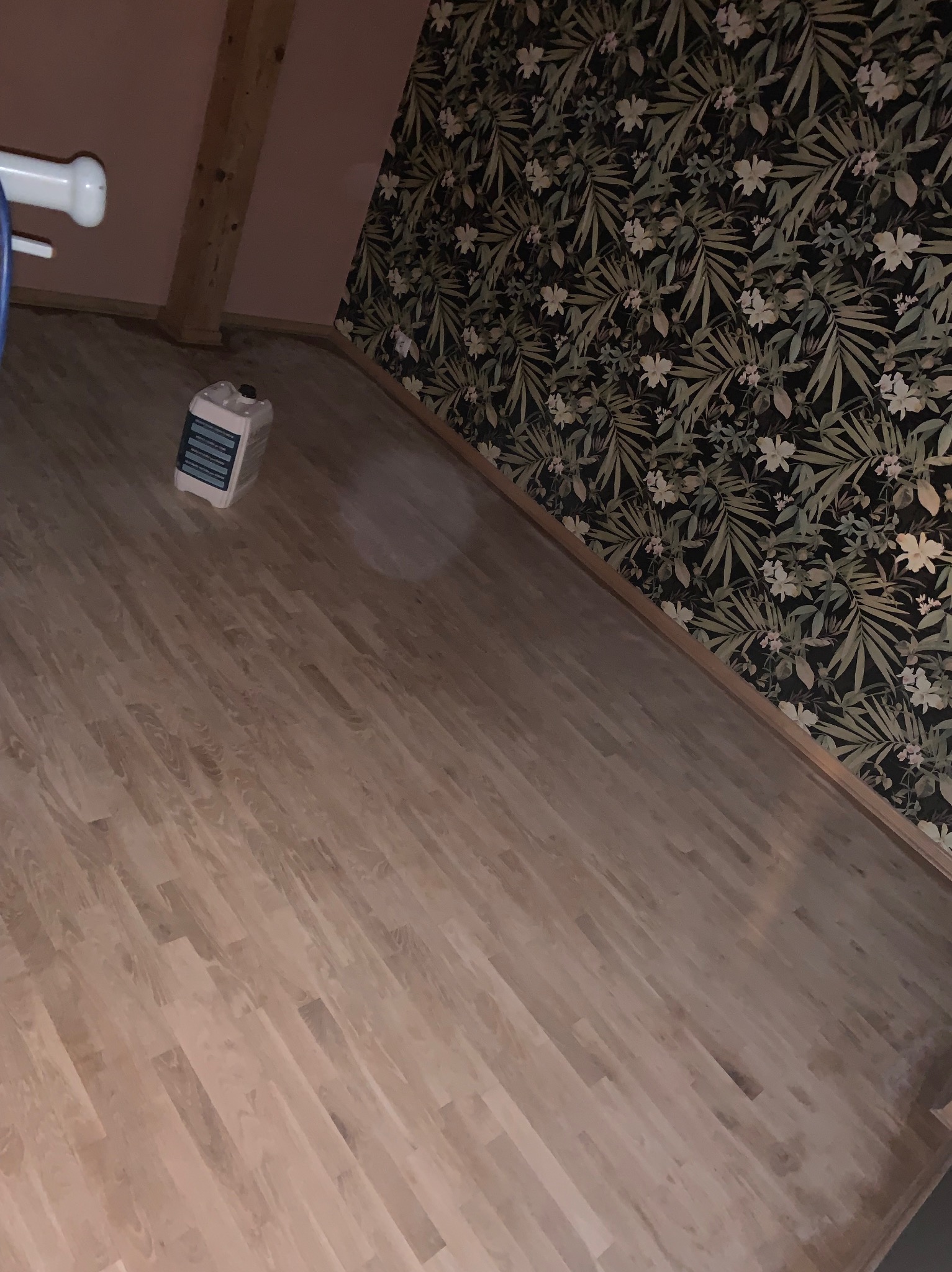Jasna, drewniana podłoga po cyklinowaniu w pokoju z ciemną tapetą w roślinny wzór. Kanister z preparatem do podłóg stoi w rogu. Widoczny fragment białego elementu.