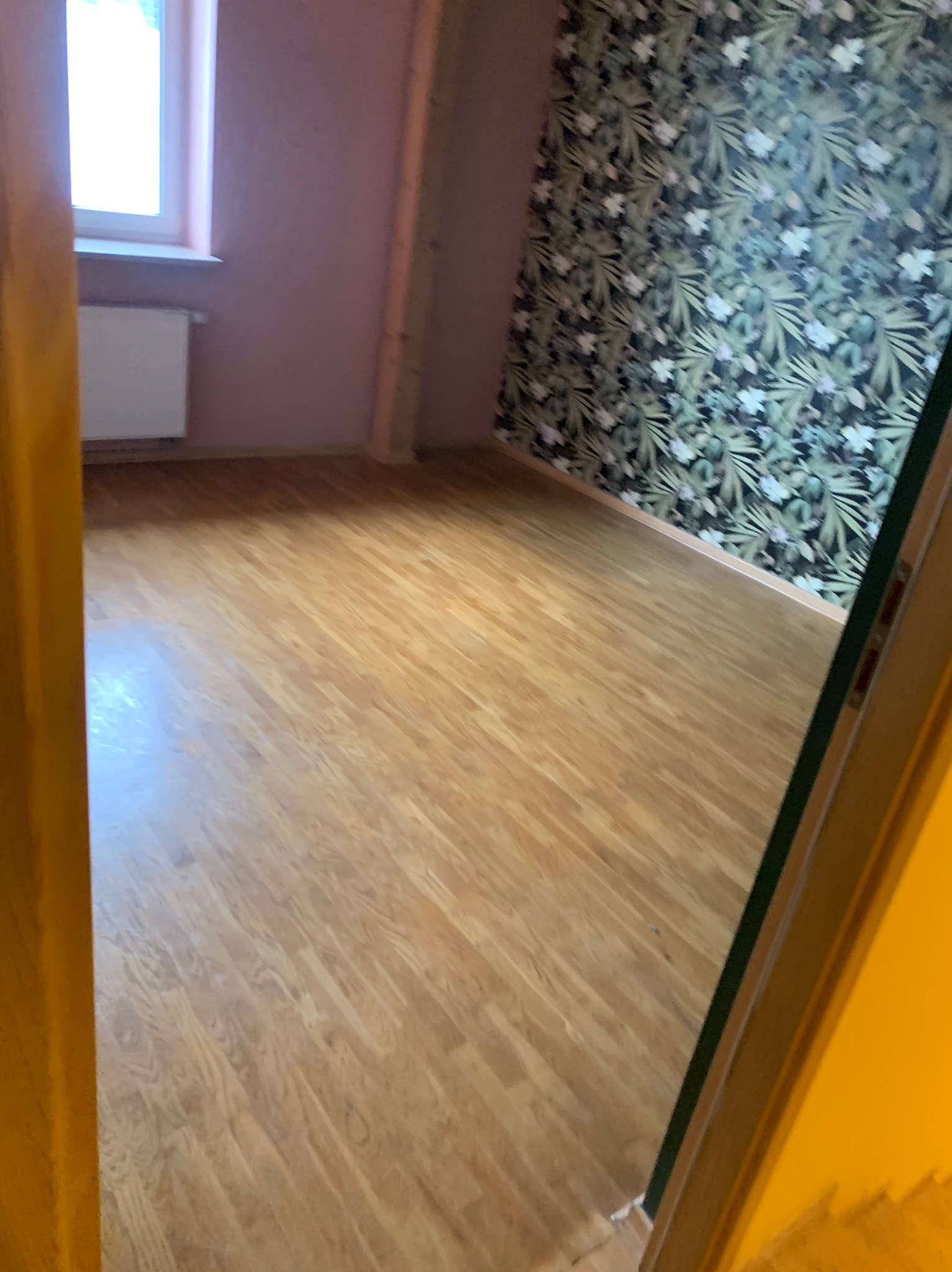 Jasna, drewniana podłoga w pokoju z różową ścianą i ścianą w zielone liście, widok zza framugi drzwi. Widoczne okno i grzejnik.