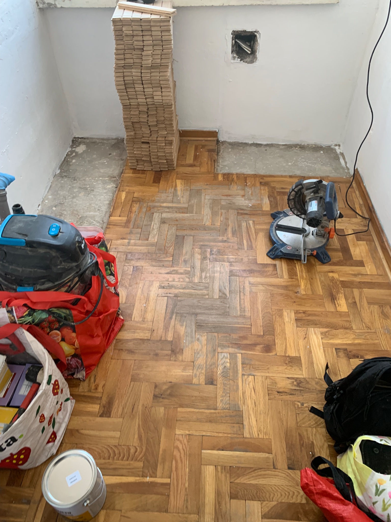 Parkiet ułożony we wzór jodełki, z narzędziami i materiałami do układania. Widoczny stos desek, piła do cięcia i odkurzacz. Częściowo przygotowane podłoże.