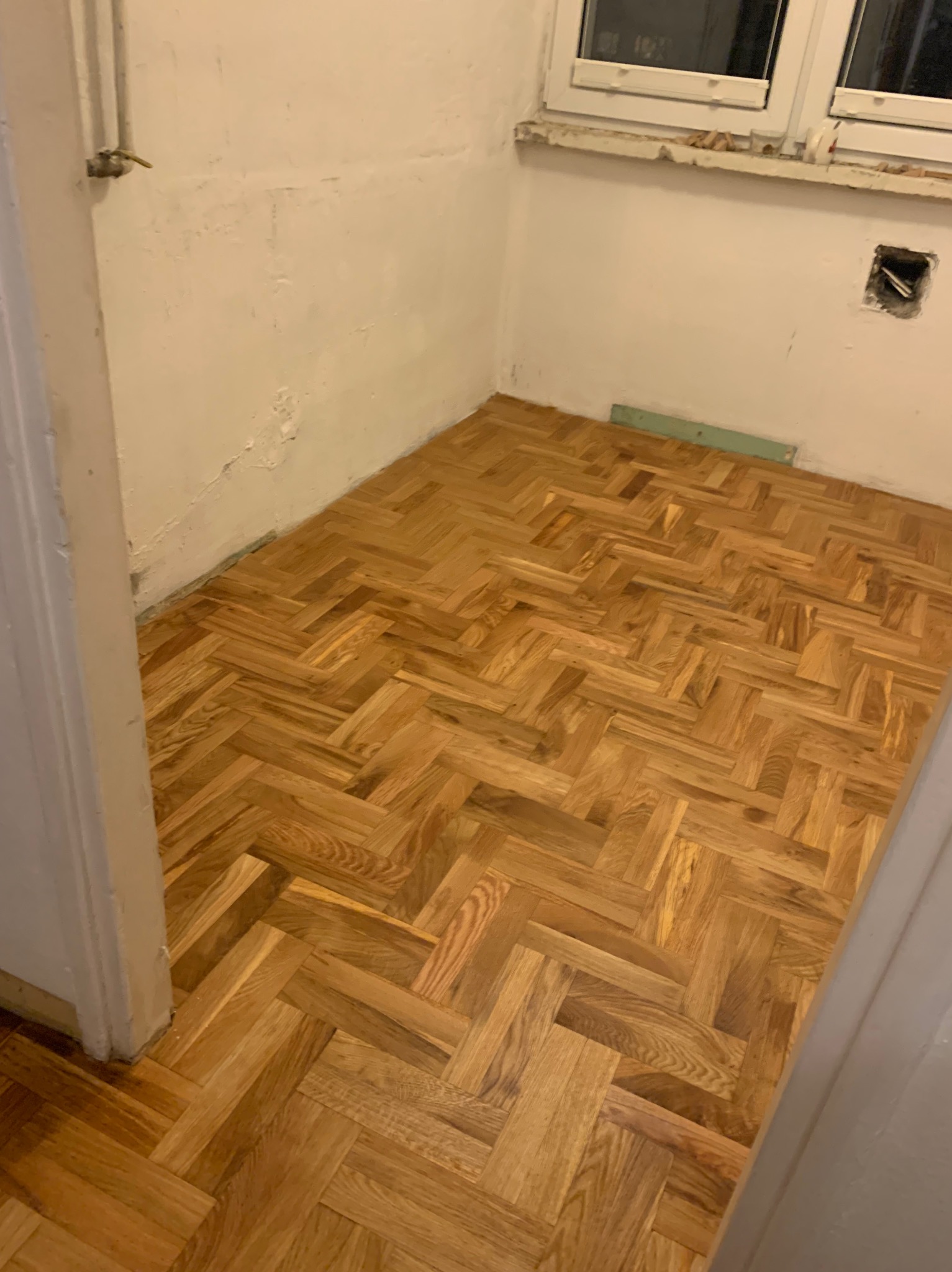 Ukończona podłoga z parkietu ułożonego w jodełkę, widoczna w rogu pomieszczenia z białymi ścianami i oknem. Fragment rury przy ścianie.