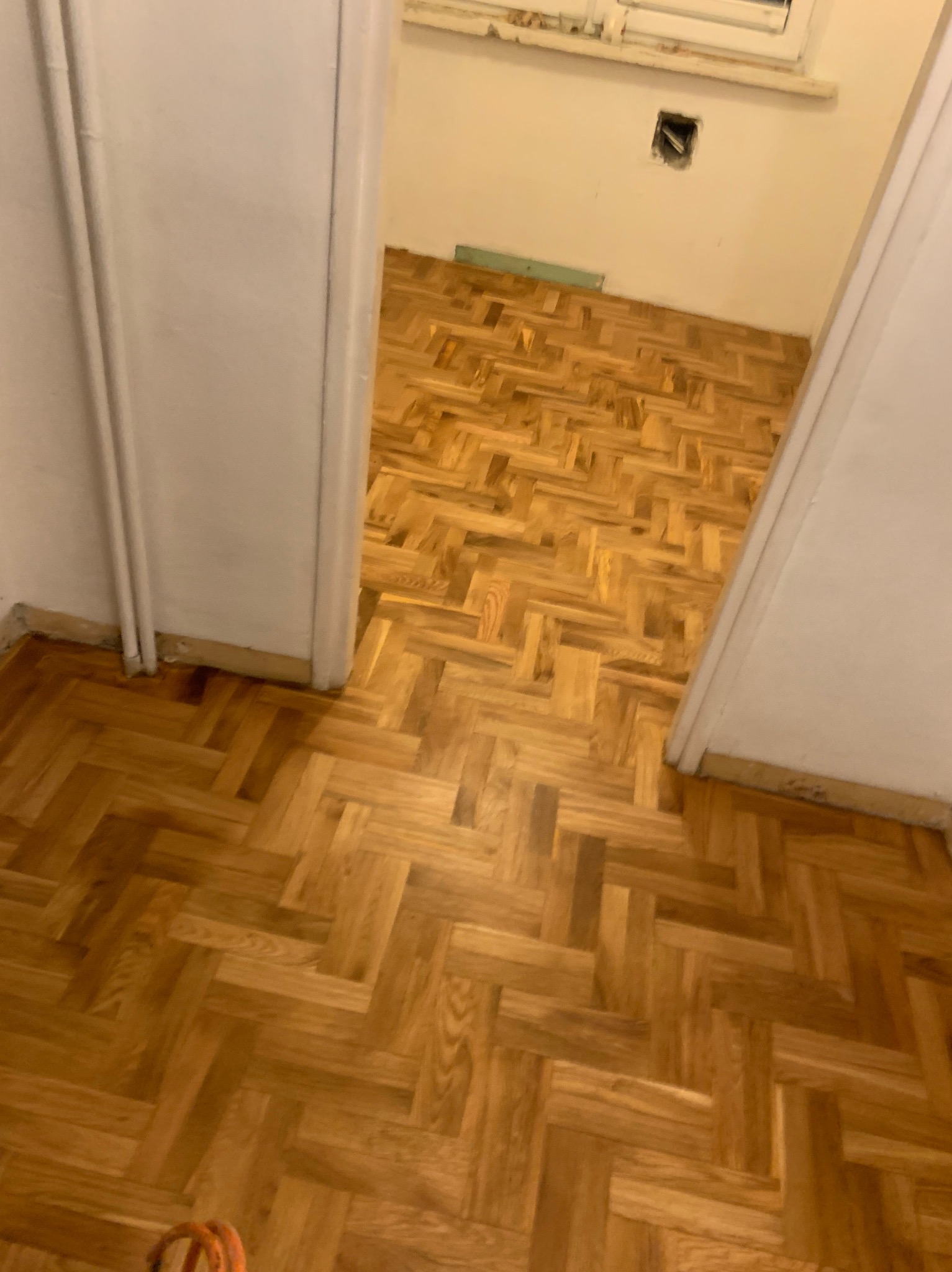 Parkiet ułożony w jodełkę, widoczny z perspektywy wejścia do pokoju. Naturalne odcienie drewna, ciepłe oświetlenie. Fragment ściany z gniazdkiem elektrycznym.