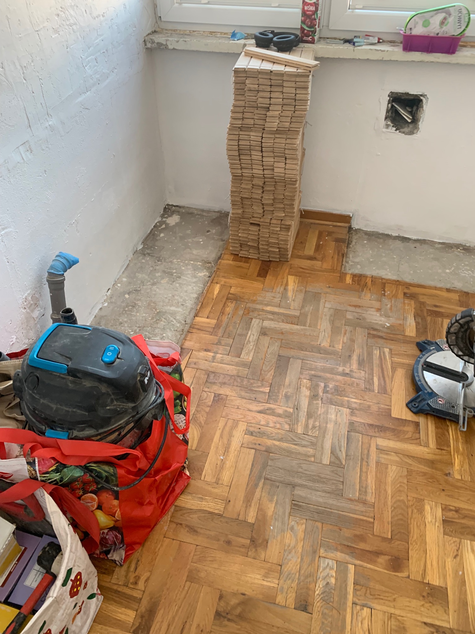 Ułożony parkiet w jodełkę, stos desek, odkurzacz budowlany i piła ukośnica w trakcie prac remontowych. Widoczne ślady po starym parkiecie.