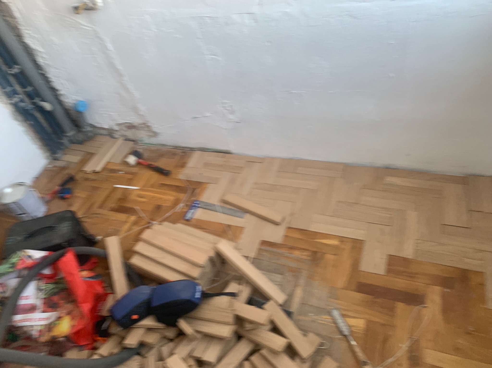 Fragment układania parkietu w jodełkę. Częściowo ułożona podłoga, narzędzia i paczki z panelami na podłodze. Widoczne rury przy ścianie.