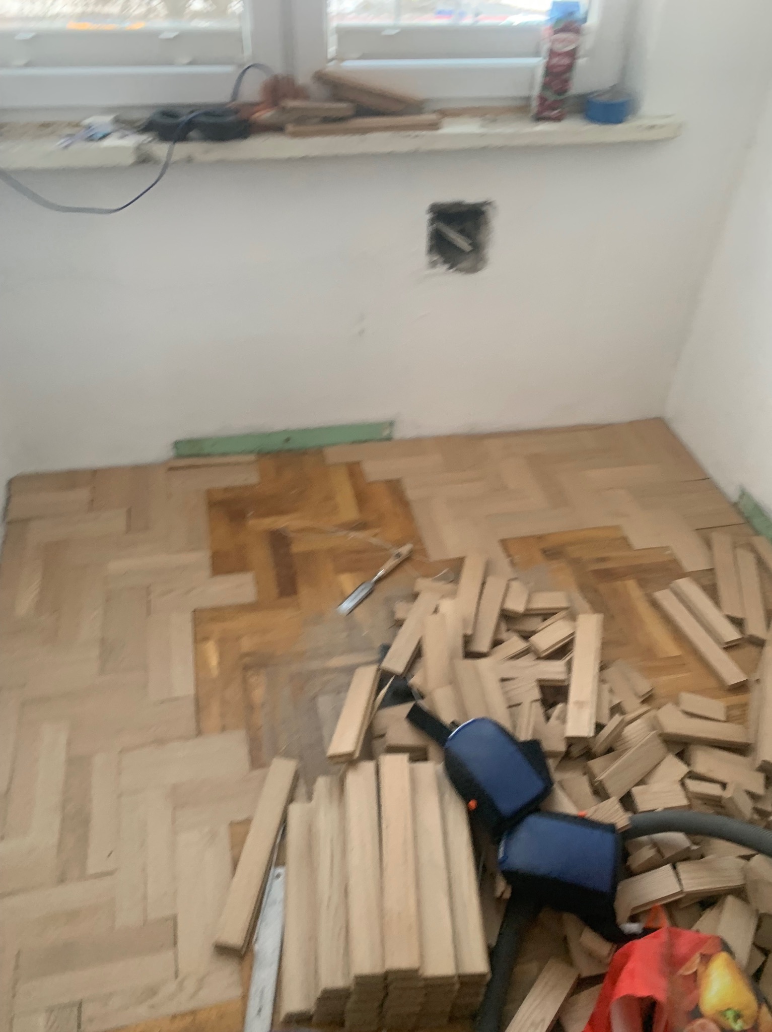 Układanie parkietu w jodełkę: częściowo ułożona podłoga, narzędzia i stosy nieułożonych klepek parkietowych w jasnym pomieszczeniu z oknem.