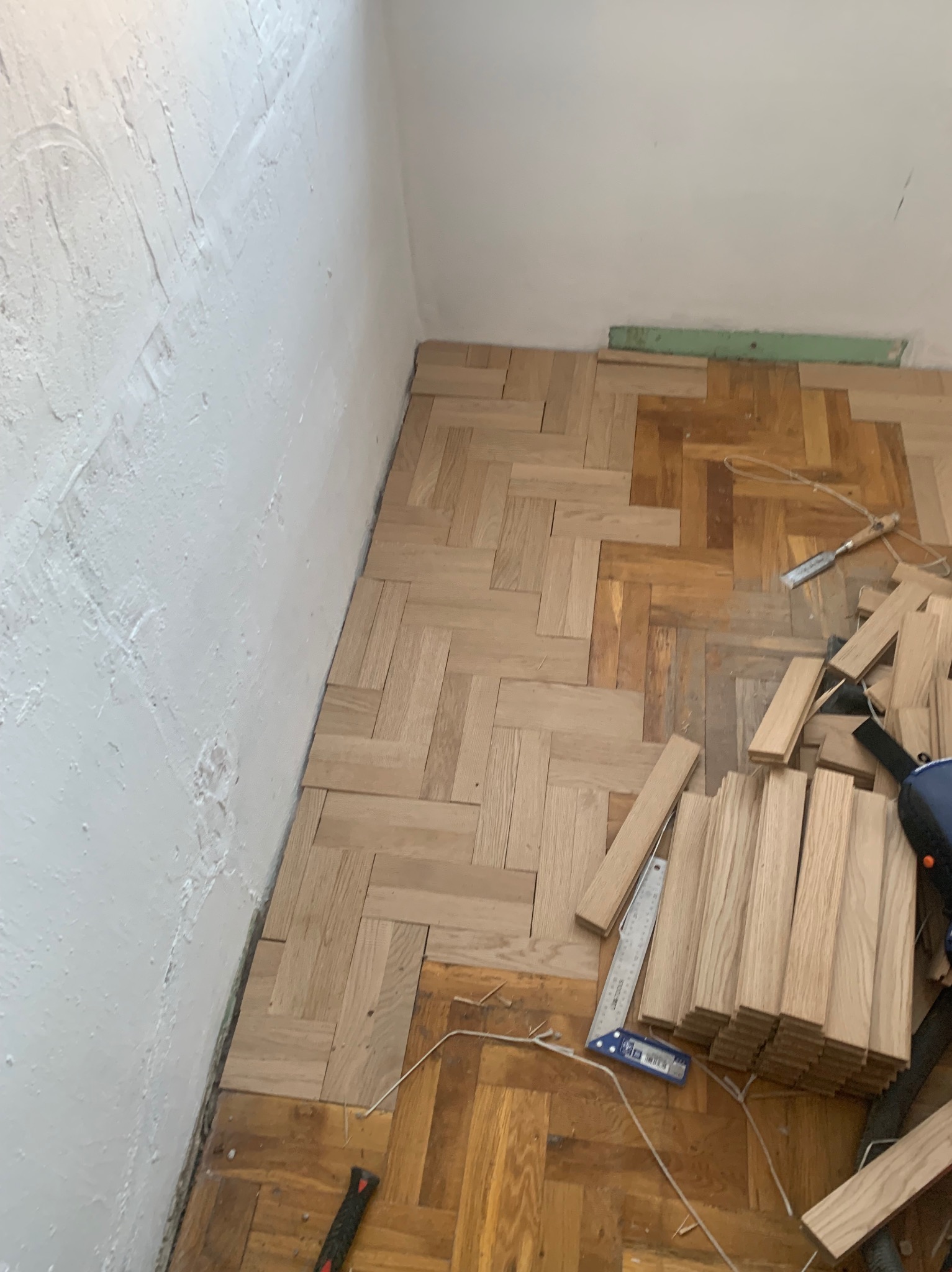 Układanie parkietu w jodełkę: fragment nowej, jasnej podłogi kontrastuje ze starą, ciemniejszą. Widoczne narzędzia i deski parkietu, ściana z nierówną fakturą.