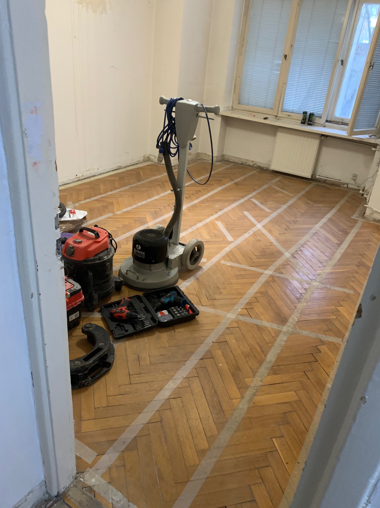 Parkiet w jodełkę przygotowany do cyklinowania, zabezpieczony taśmą. Widoczna cykliniarka, odkurzacz przemysłowy i narzędzia. Ujęcie zza progu.