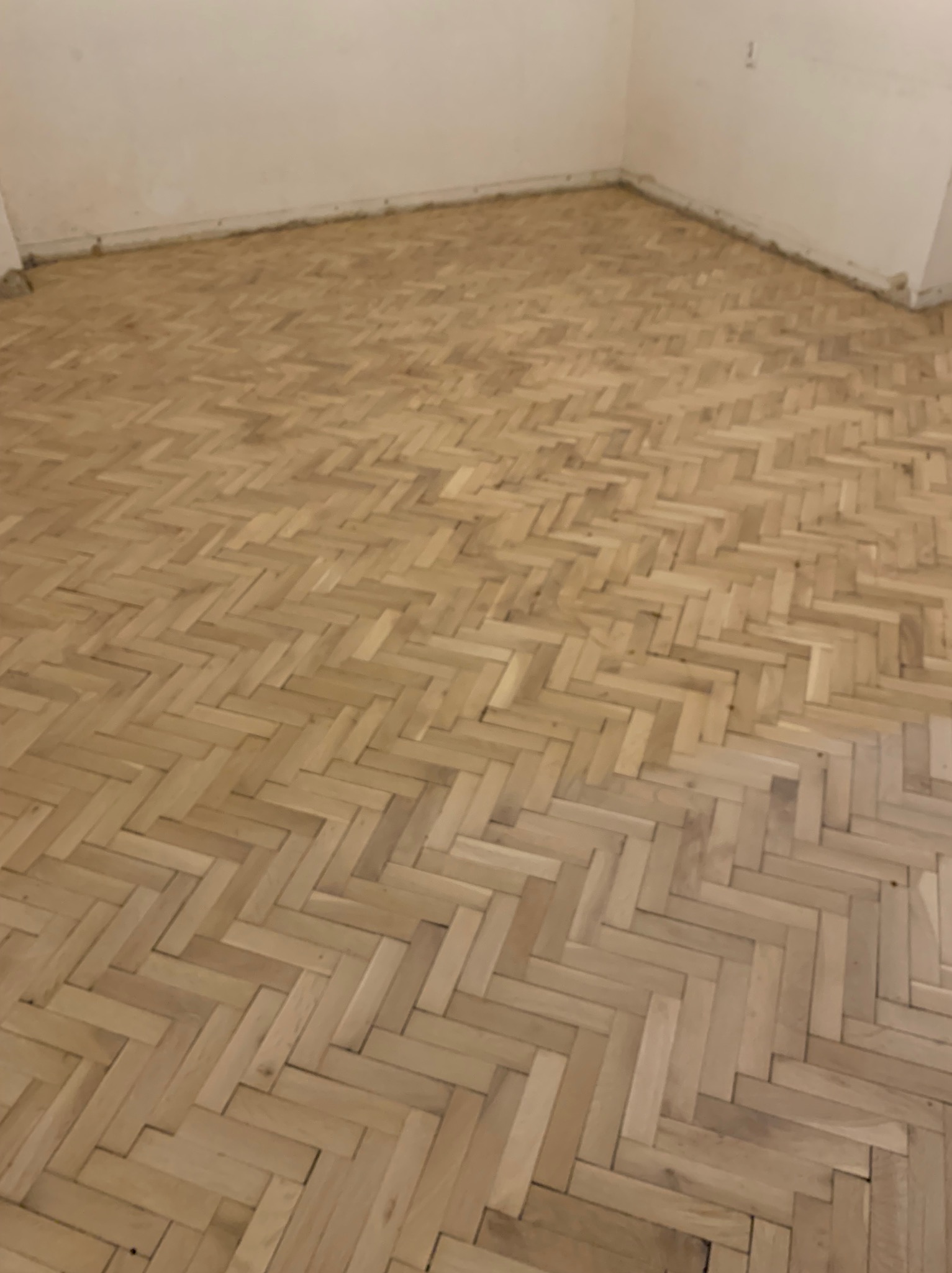 Jasny, odnowiony parkiet ułożony w jodełkę w pustym pokoju z białymi ścianami. Widoczne ślady po listwach przypodłogowych. Perspektywa z narożnika pomieszczenia.