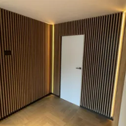 Nowoczesne wnętrze z drewnianą ścianą z lameli, oświetleniem LED i parkietem w jodełkę, białe drzwi z czarnymi klamkami.