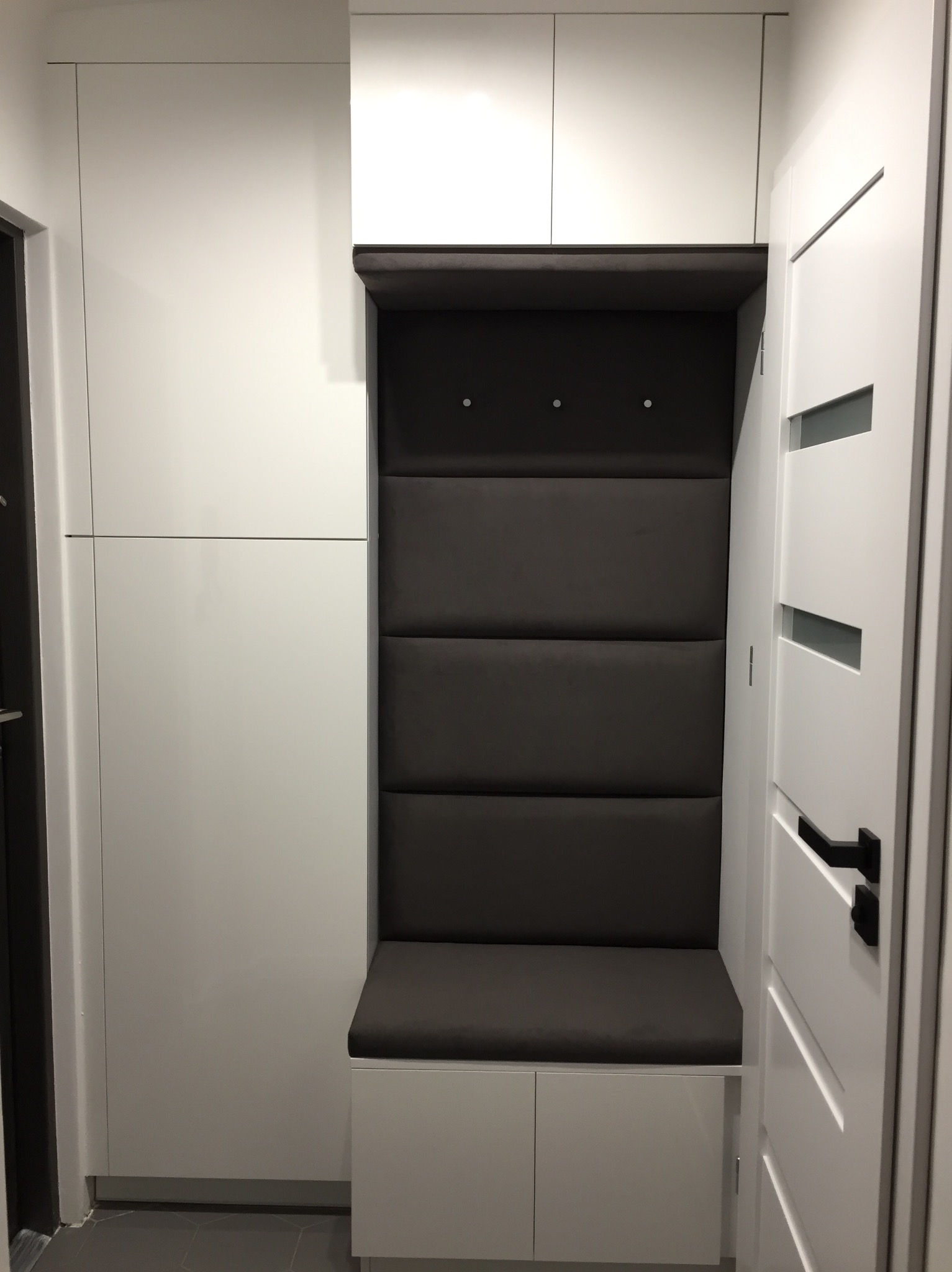 Wnęka z siedziskiem obita szarą tapicerką, z trzema wieszakami, wkomponowana w białą zabudowę meblową w przedpokoju, obok białe drzwi z czarną klamką.