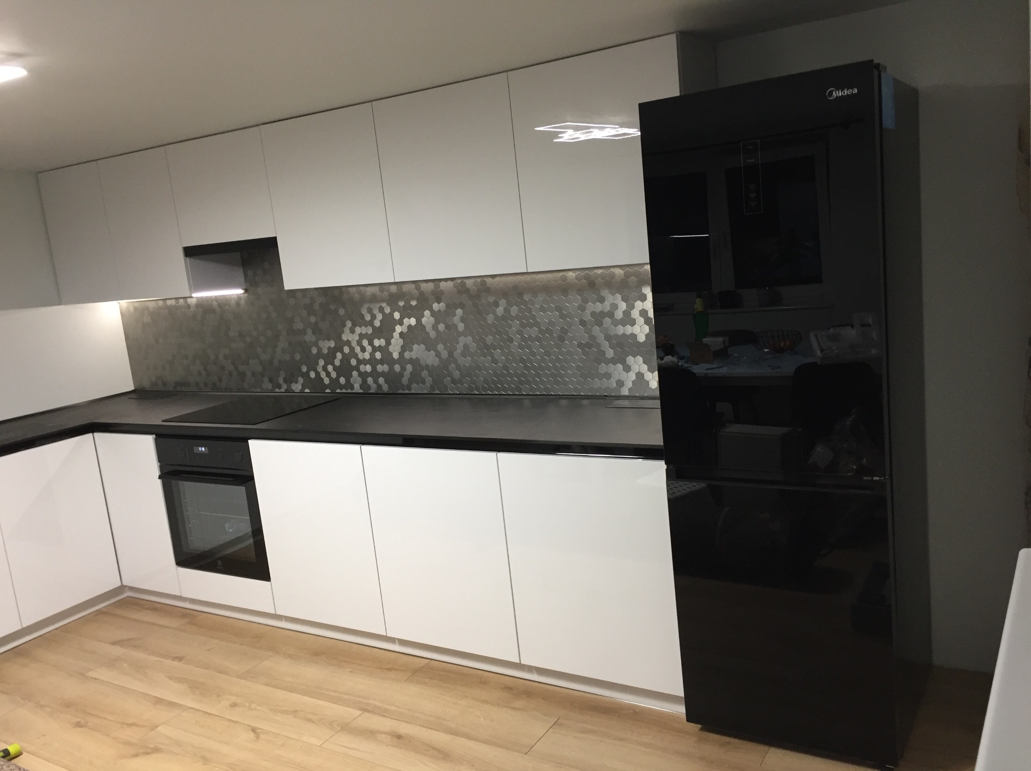 Nowoczesna kuchnia z białymi, lakierowanymi frontami szafek, czarnym blatem i lodówką Midea; ściana nad blatem wykończona mozaiką w kształcie plastra miodu.