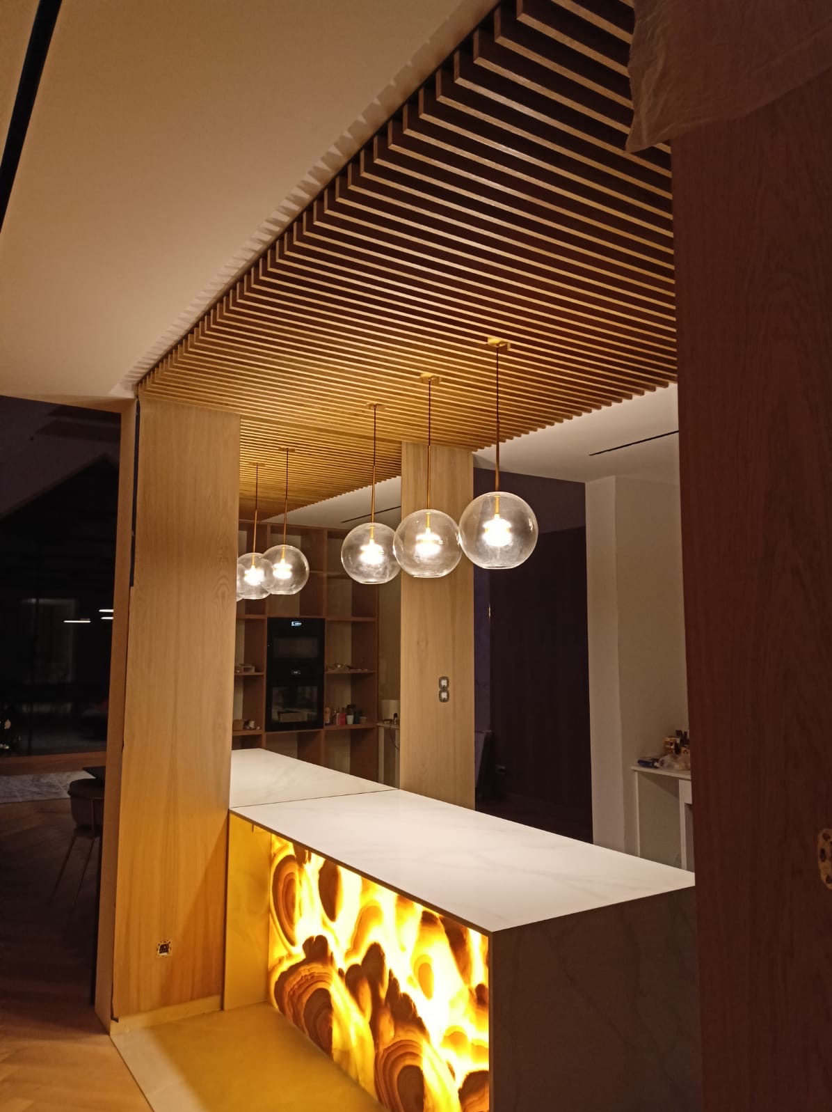 Nowoczesna kuchnia z drewnianym sufitem lamelowym, oświetleniem kulistymi lampami wiszącymi i wyspą z podświetlanym frontem z onyksu.