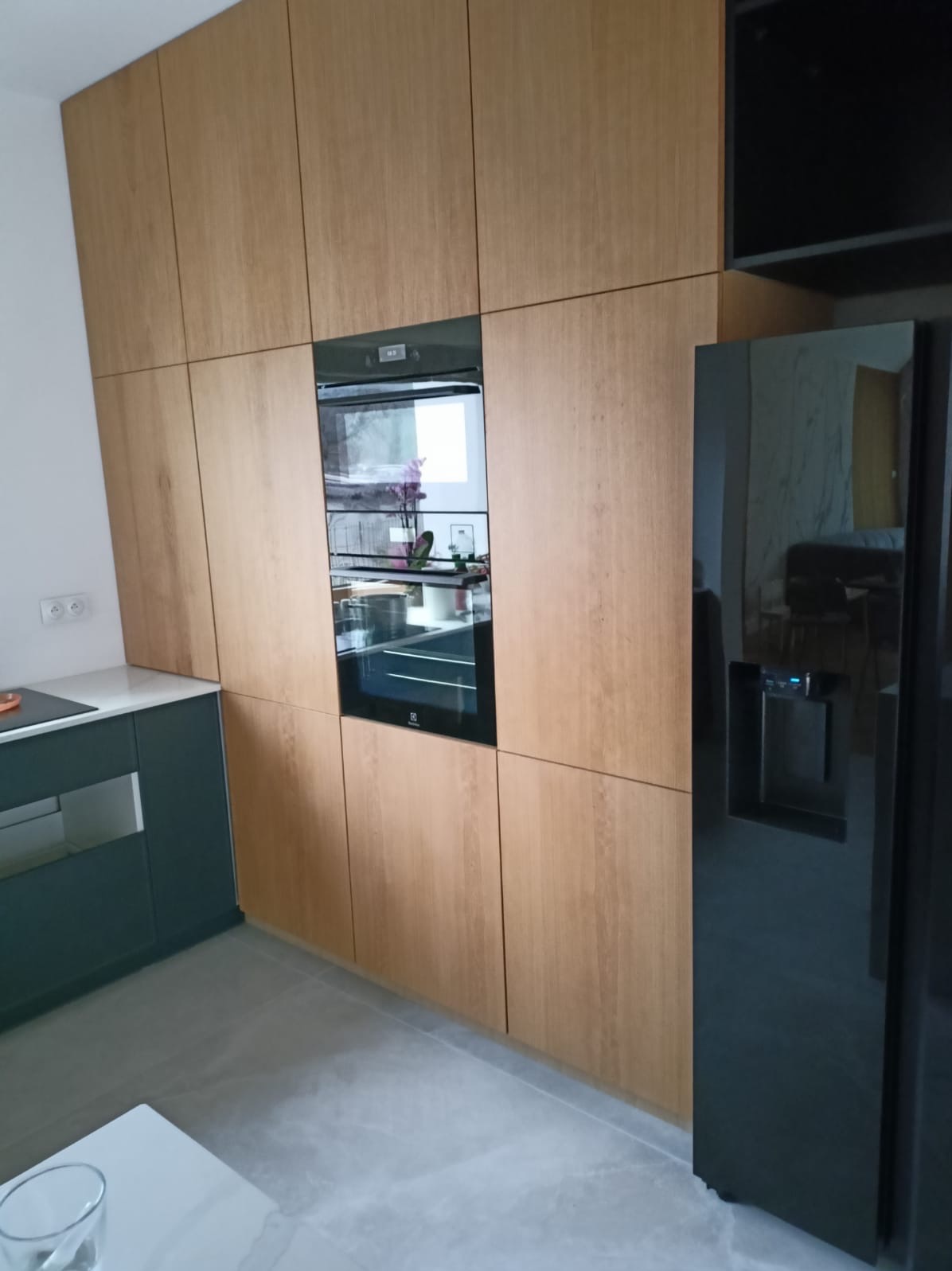 Nowoczesna kuchnia z drewnianymi frontami, wbudowanym piekarnikiem i czarną lodówką side-by-side z dystrybutorem wody, widok z dołu, szare płytki podłogowe.