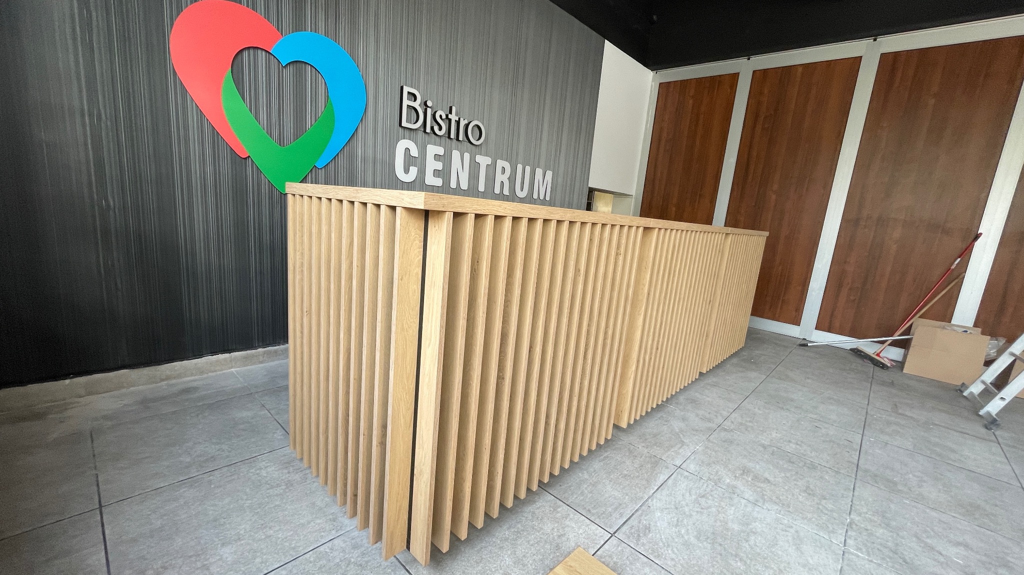 Lamelowa lada recepcyjna z jasnego drewna, wykonana na wymiar w Dobroniu, z widocznym logo 'Bistro Centrum' w tle na grafitowej ścianie.