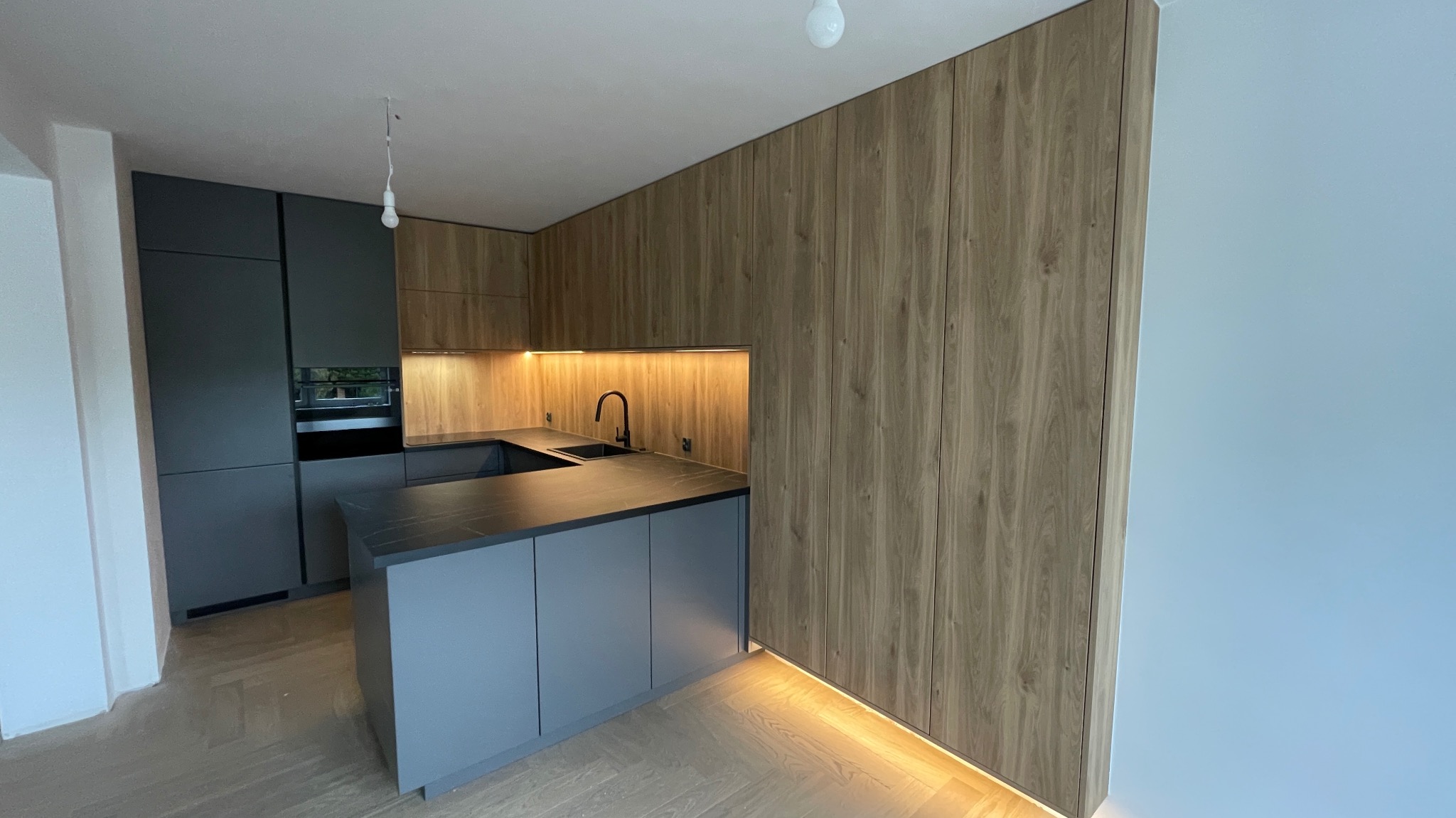 Nowoczesna kuchnia z szarymi szafkami i drewnianą okładziną ścienną, podświetleniem LED pod szafkami i blatem z czarnego kamienia.