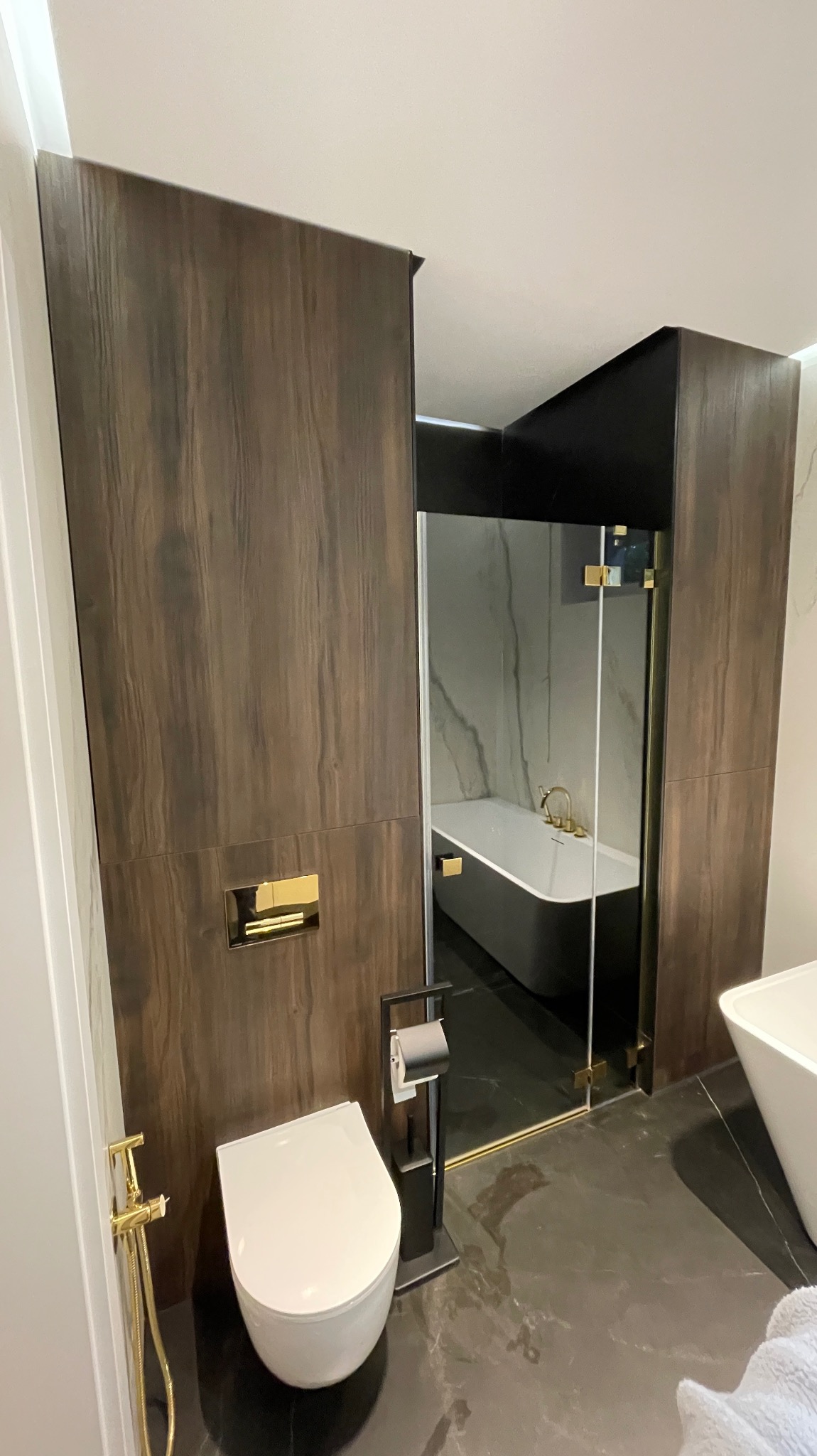 Nowoczesna łazienka z ciemnymi panelami imitującymi drewno, złotą armaturą, wiszącą toaletą i wolnostojącą wanną widoczną przez przeszkloną ściankę.