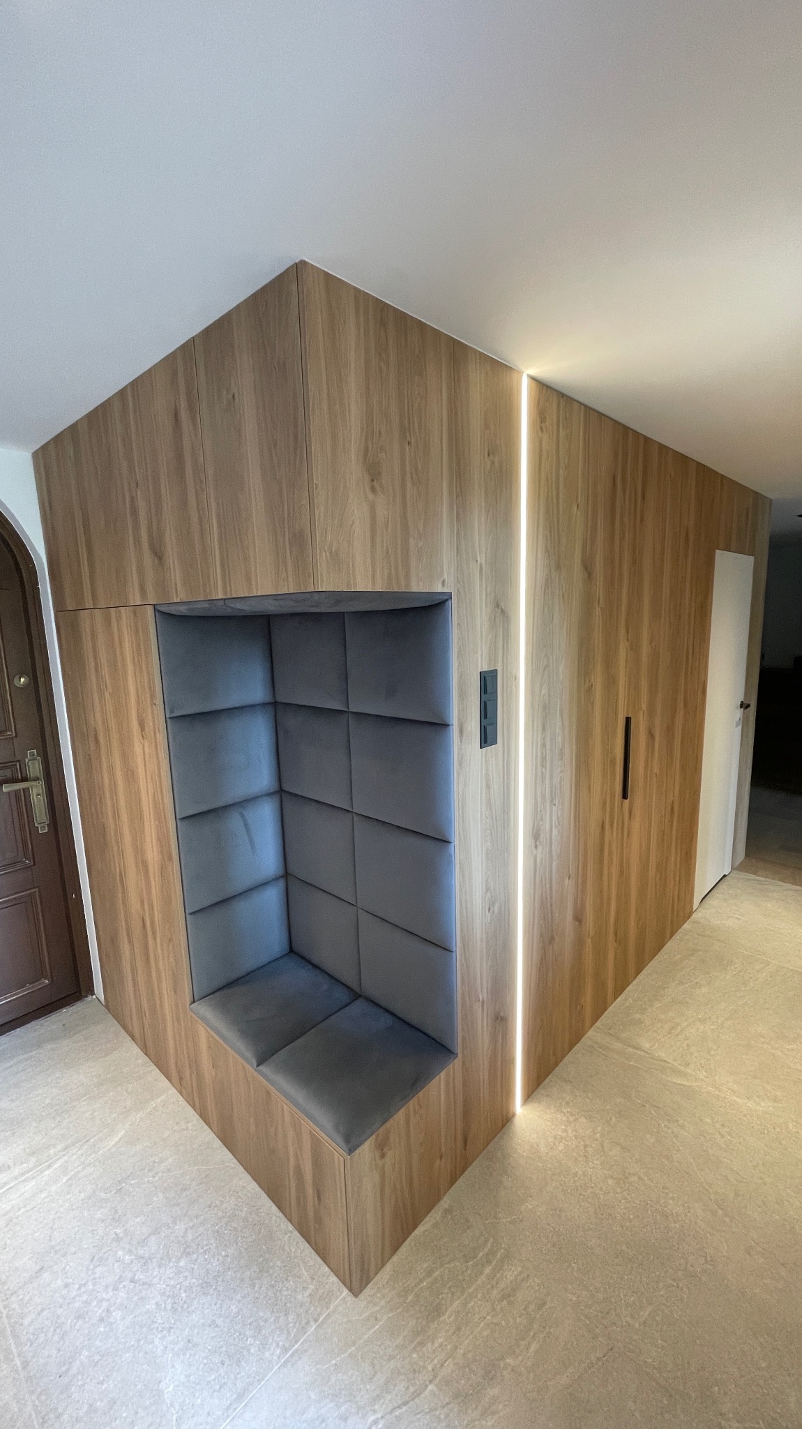 Wnęka z szarą tapicerką w drewnianej zabudowie, zintegrowana z oświetleniem LED wzdłuż ściany, widok na przedpokój z drzwiami wejściowymi.