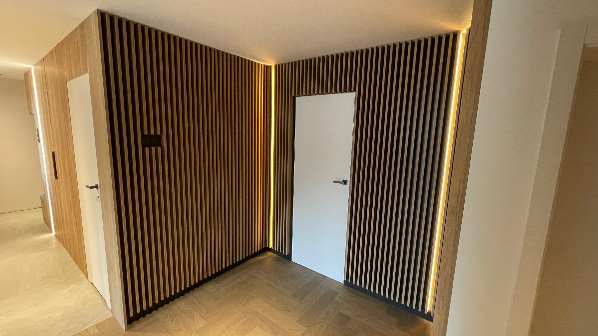 Nowoczesne wnętrze z drewnianą ścianą z lameli, oświetleniem LED i parkietem w jodełkę, białe drzwi z czarnymi klamkami.