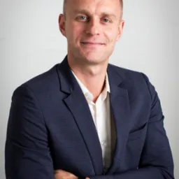 Uśmiechnięty mężczyzna w granatowej marynarce i białej koszuli, z zegarkiem na ręku, pozuje na jasnym tle. Profesjonalny wizerunek.