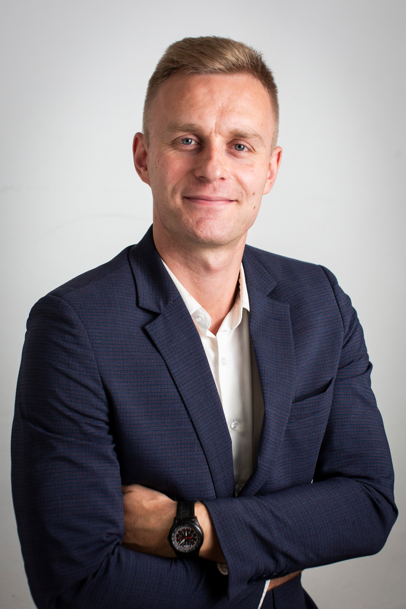 Uśmiechnięty mężczyzna w granatowej marynarce i białej koszuli, z zegarkiem na ręku, pozuje na jasnym tle. Profesjonalny wizerunek.