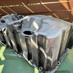 Piotr Kryś - Oczyszczona, aluminiowa miska olejowa silnika samochodowego, widoczne ślady użycia, leżąca na zielonym koszu na śmieci, w tle drewniany płot.