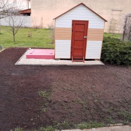 Piotr Kryś - Kolorowa, drewniana budka narzędziowa z czerwoną klapą do piwnicy, otoczona świeżo przekopaną ziemią i kamieniami w ogrodzie.