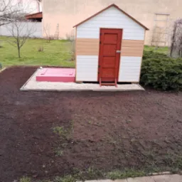 Kolorowa, drewniana budka narzędziowa z czerwoną klapą do piwnicy, otoczona świeżo przekopaną ziemią i kamieniami w ogrodzie.