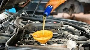 Napełnianie silnika olejem przez pomarańczową dłoń w rękawicy, widok na silnik samochodowy z lejkiem i butelką oleju, warsztatowe otoczenie.