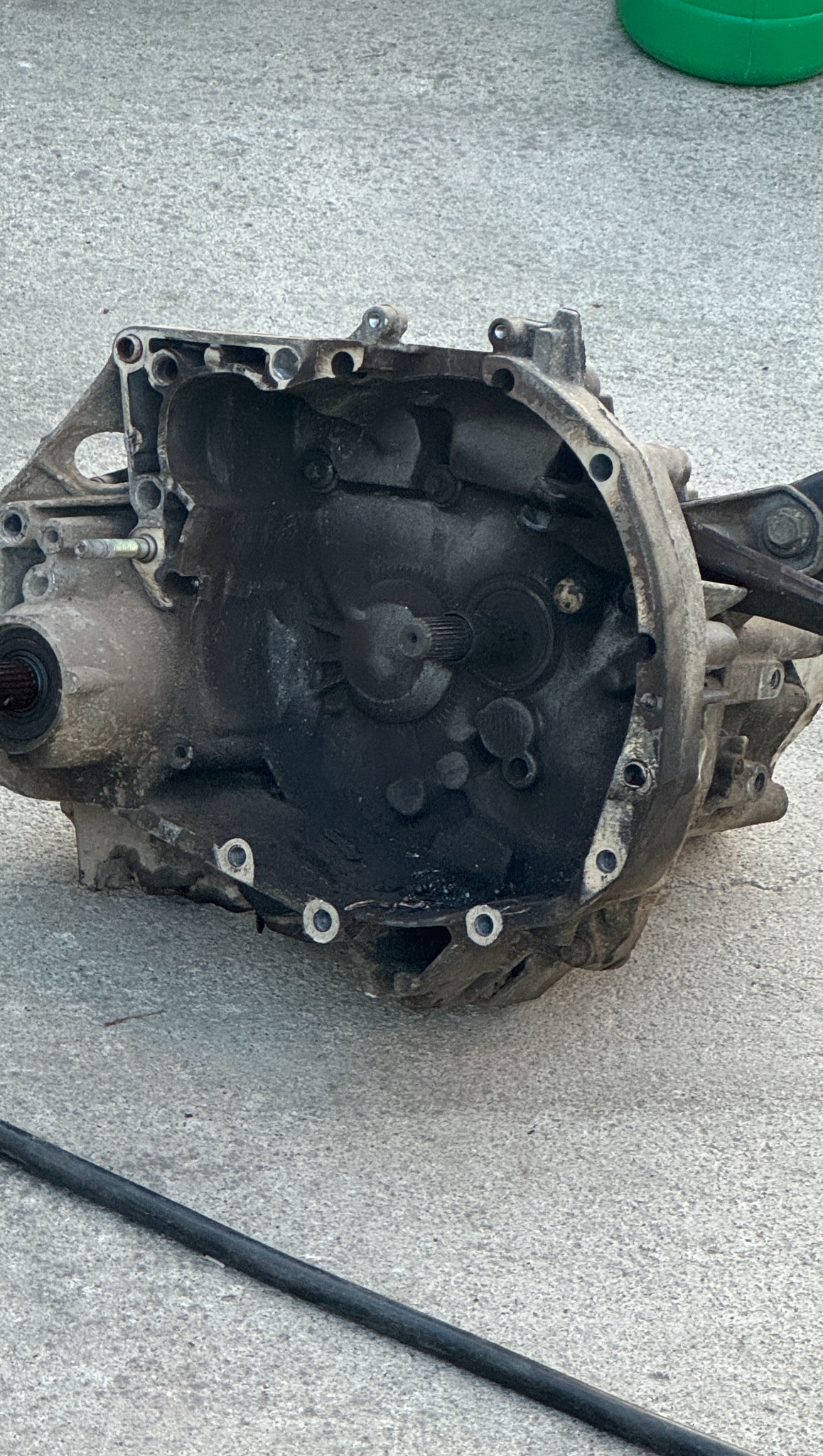 Brudna, zdemontowana skrzynia biegów na szarym tle betonowym. Widoczne ślady oleju i zużycia. Otwarta obudowa ukazuje wewnętrzne mechanizmy przekładni.