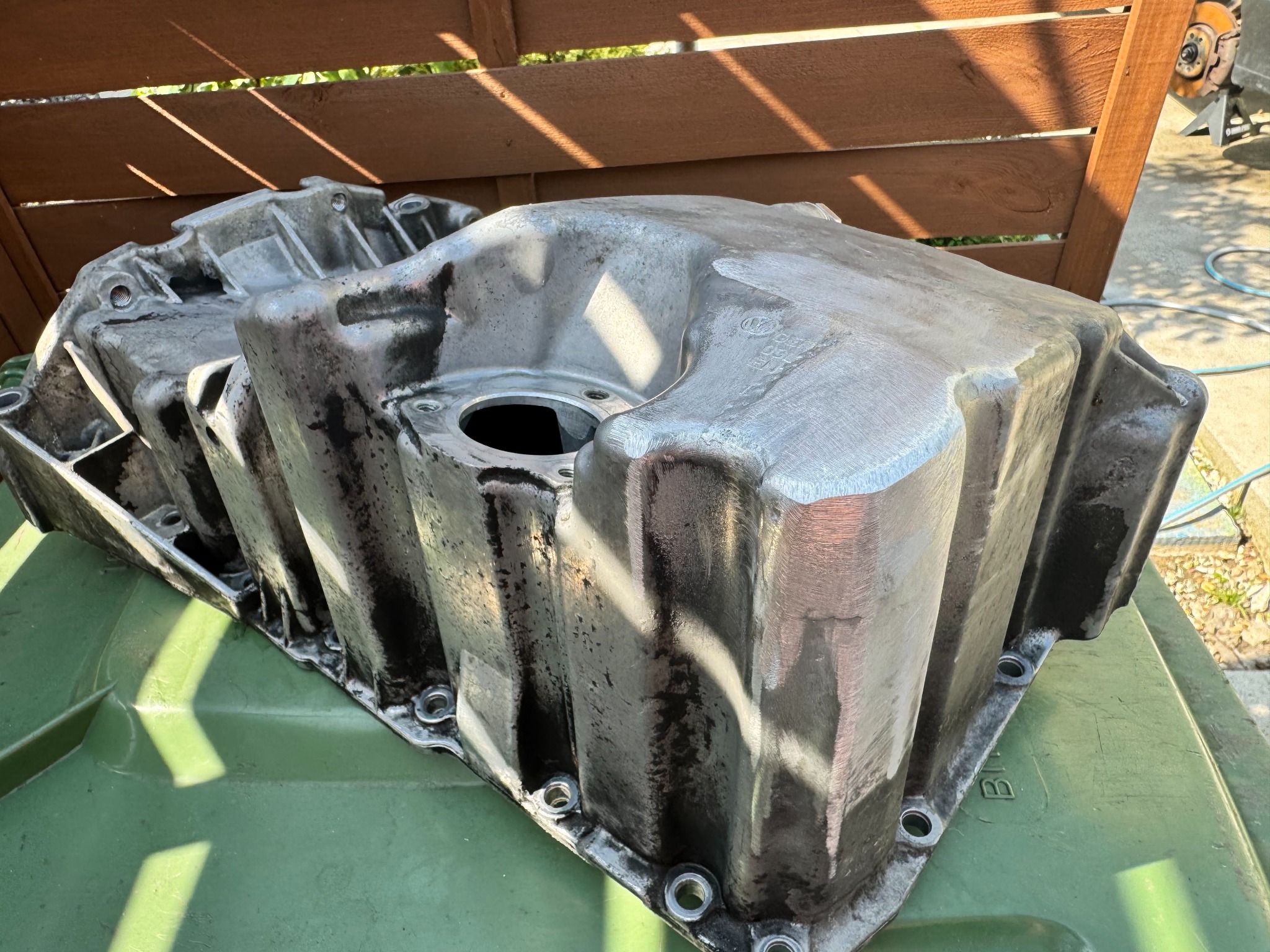 Oczyszczona, aluminiowa miska olejowa silnika samochodowego, widoczne ślady użycia, leżąca na zielonym koszu na śmieci, w tle drewniany płot.