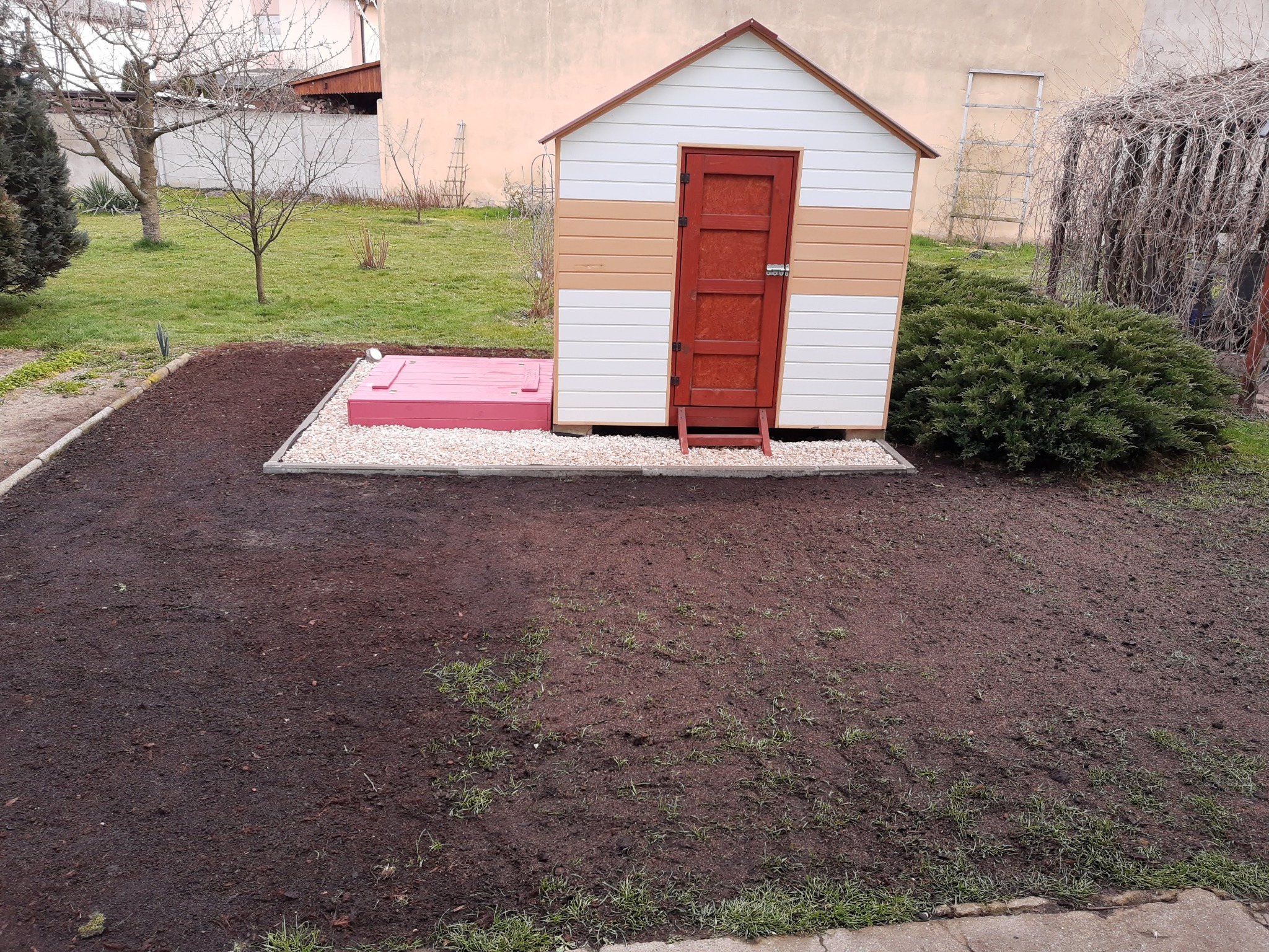 Kolorowa, drewniana budka narzędziowa z czerwoną klapą do piwnicy, otoczona świeżo przekopaną ziemią i kamieniami w ogrodzie.