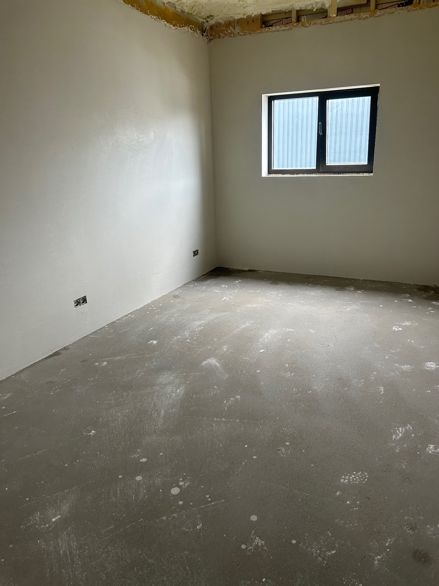 Surowe, jasne wnętrze po nałożeniu gładzi gipsowej na ściany, z widocznymi puszkami elektrycznymi i oknem z żaluzjami.